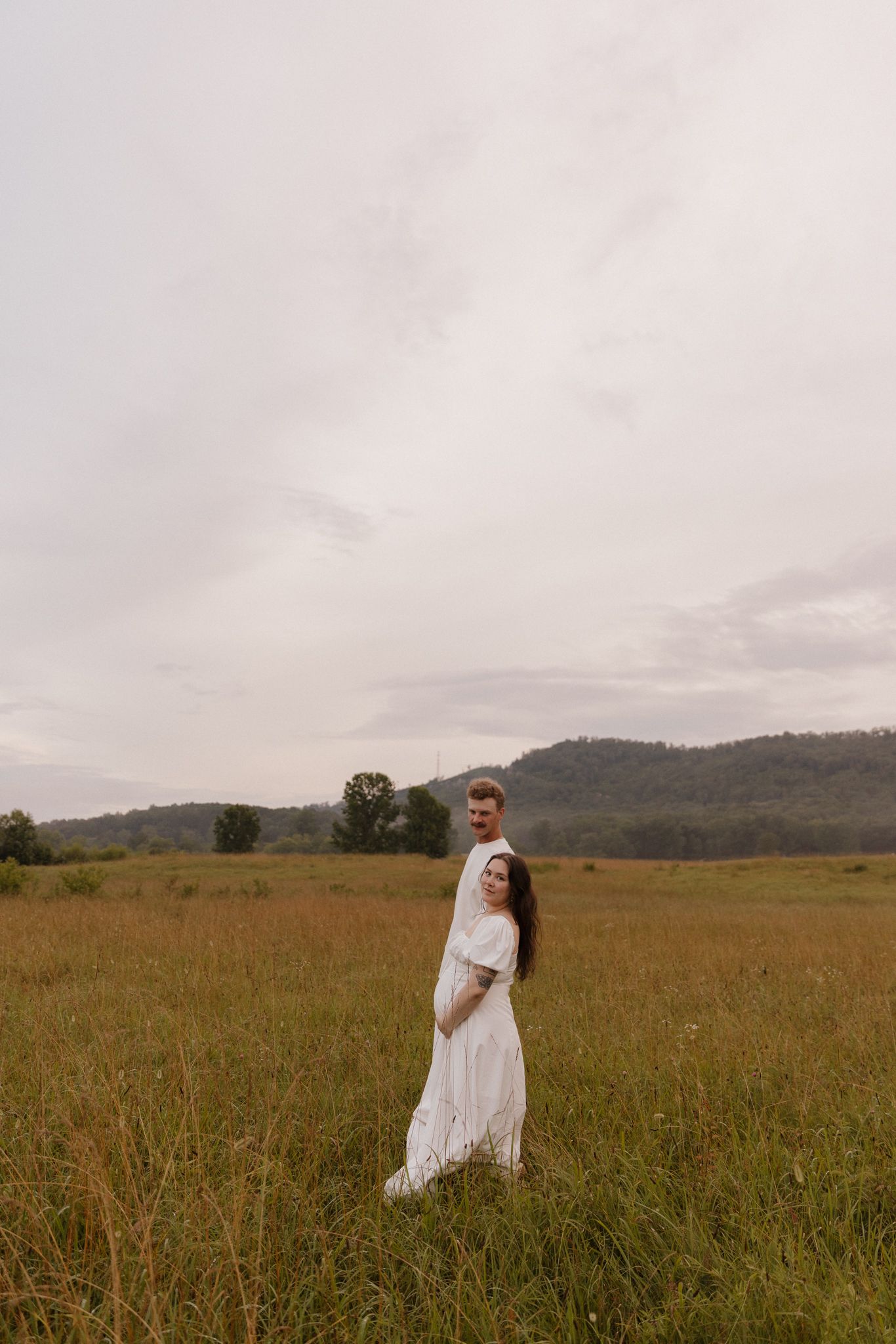 A woman in a white dress stands in a golden field with mountains visible in the background during sunset.