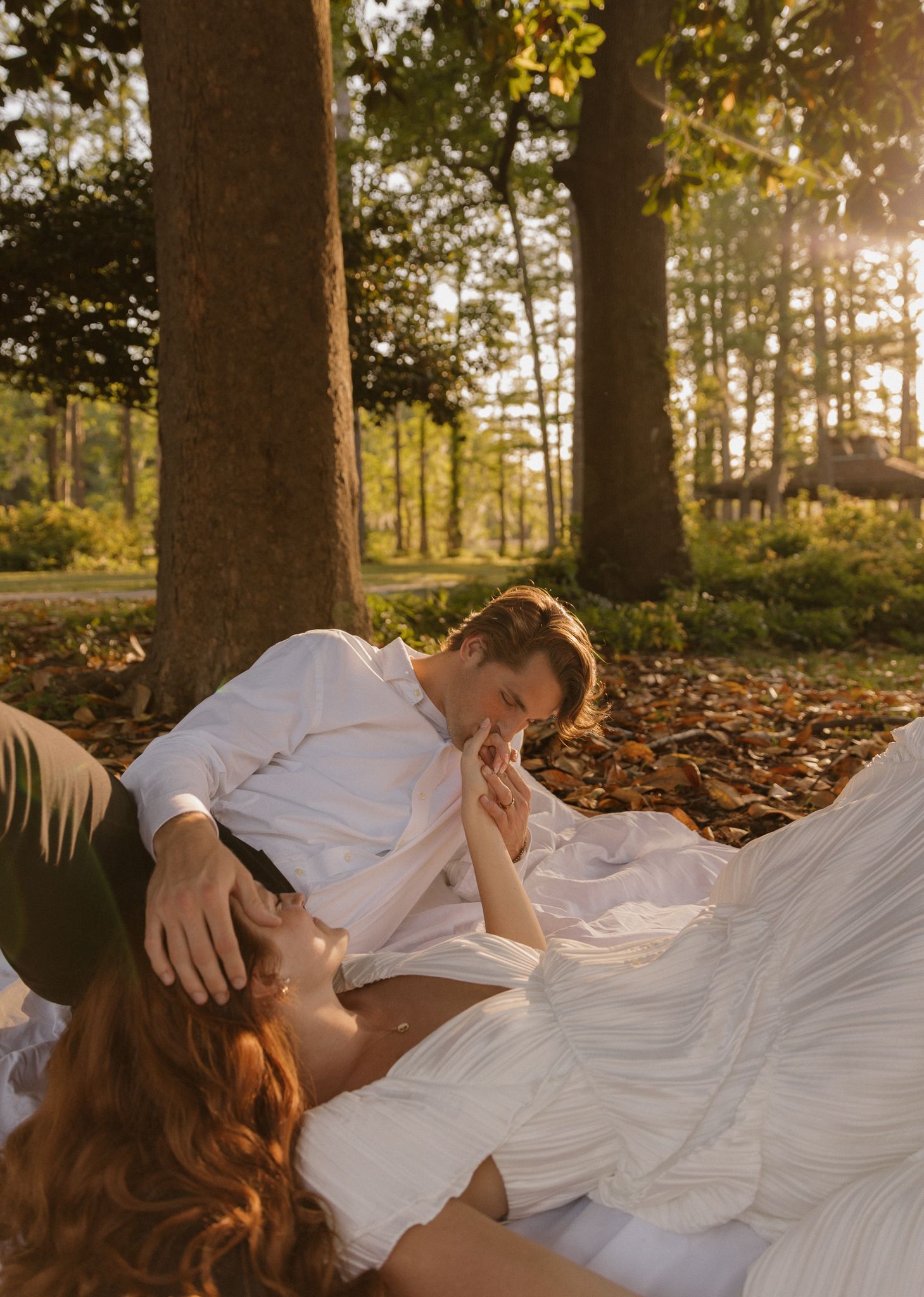 A romantic couple in white clothing lying on autumn leaves in a forest during golden hour sunset.