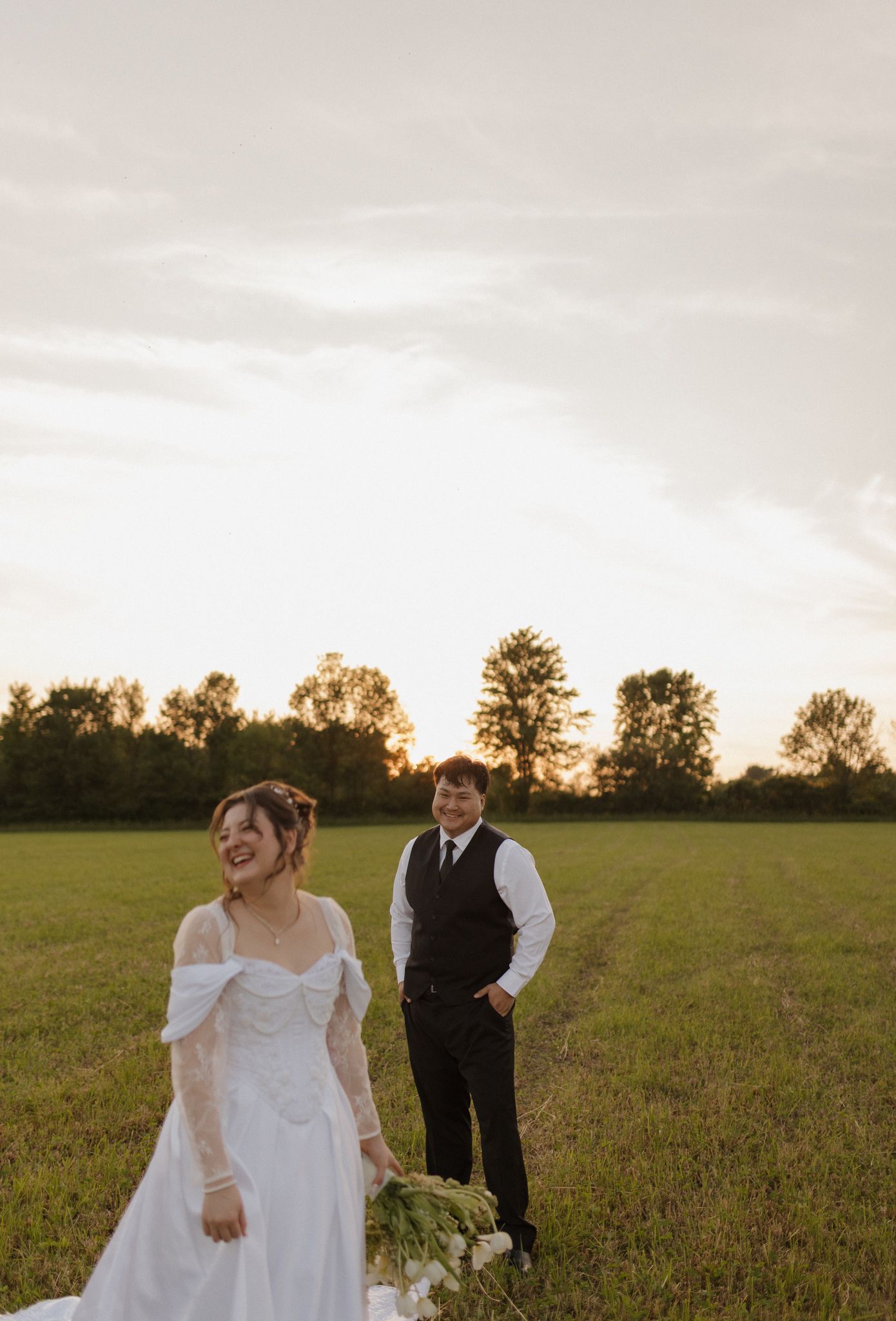 A wedding couple stands in a golden field at sunset with trees silhouetted in the background.