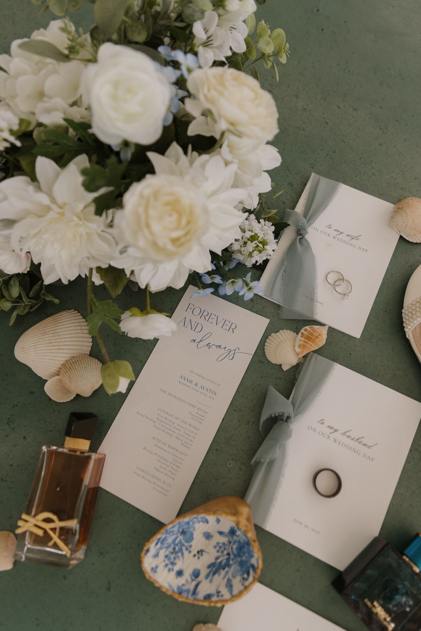 Wedding detail flatlay with white flowers, invitation cards, and jewelry on a green surface.