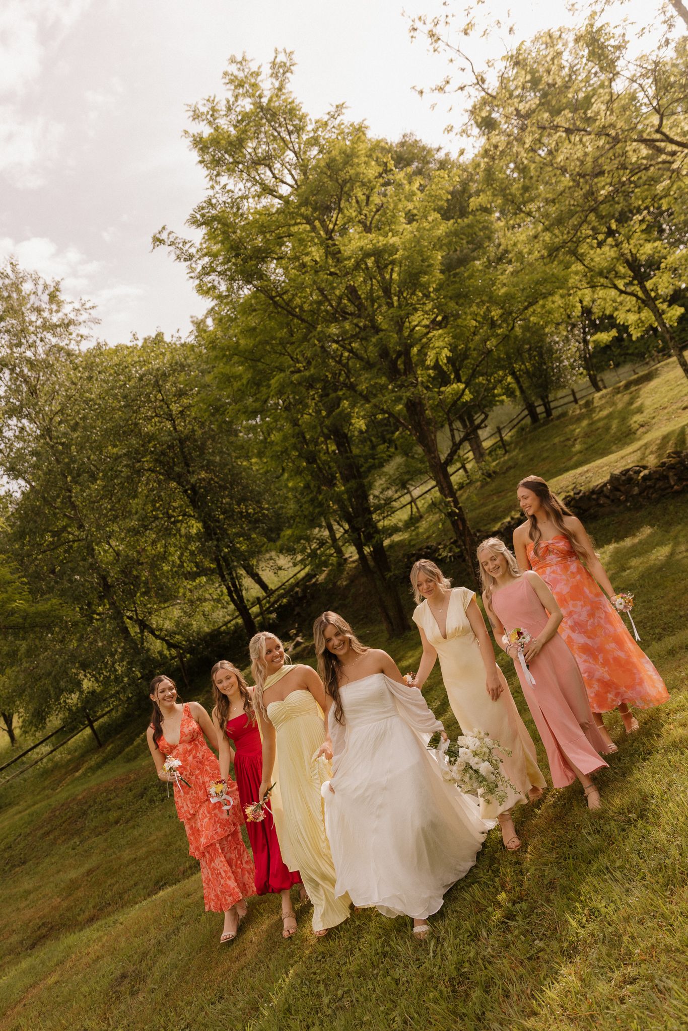 Wedding party in coral and yellow dresses walking through a grassy hillside at sunset.