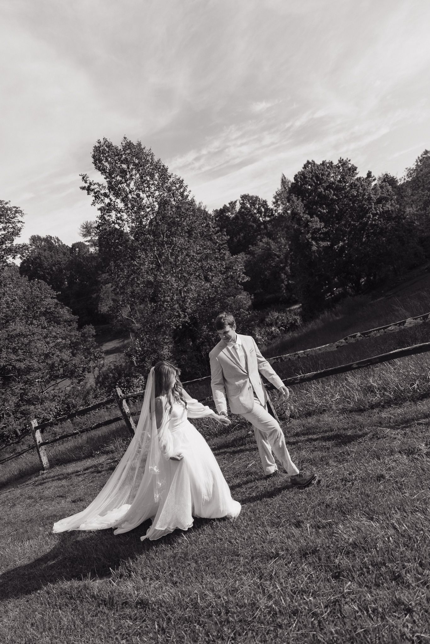 Black and white wedding photo of couple walking along rustic fence line in countryside.