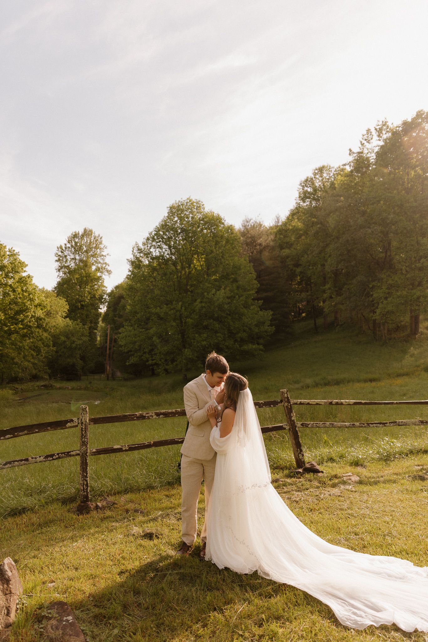 Wedding couple shares intimate moment on grassy hillside with long cathedral veil trailing behind.