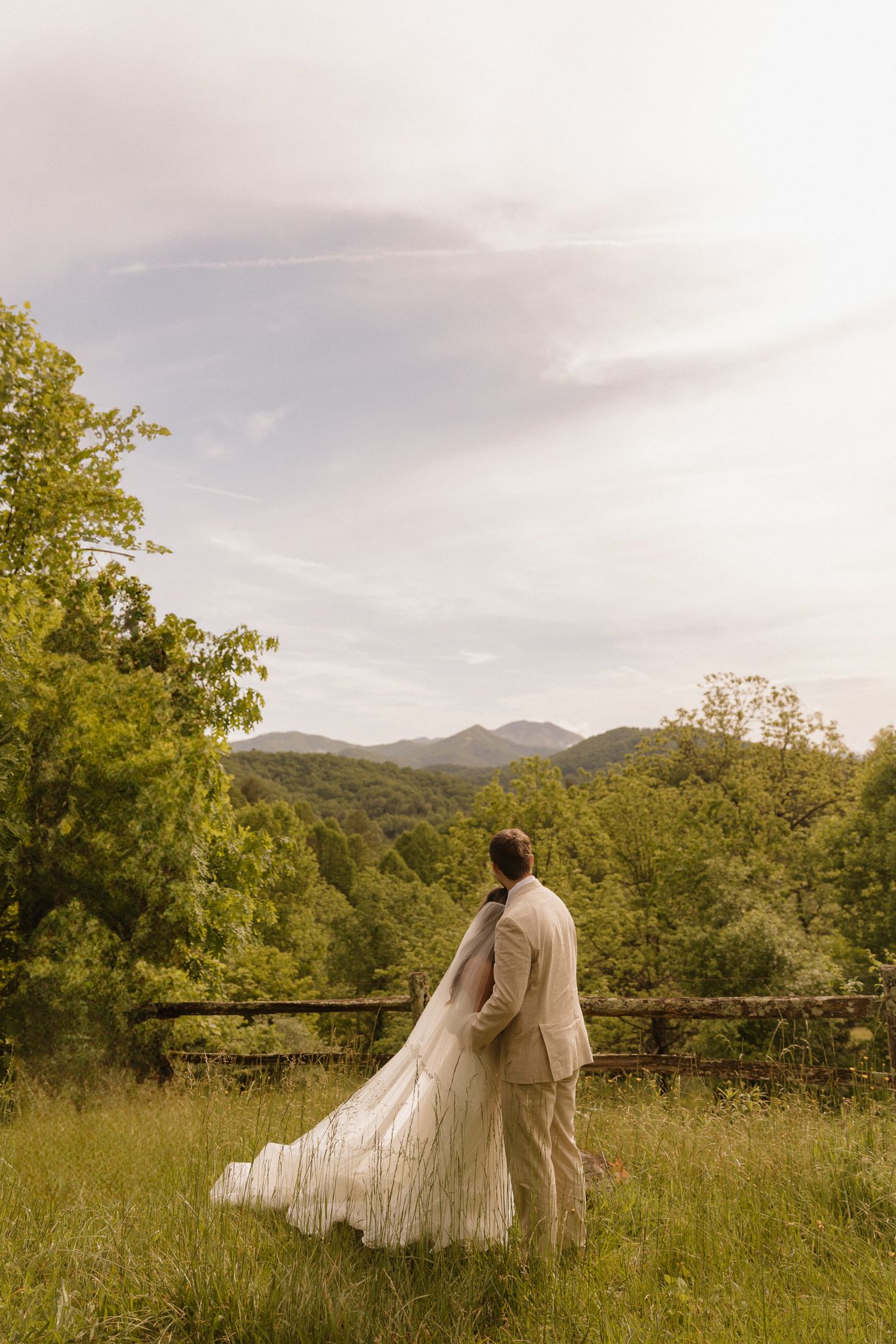 Couple in wedding attire walking through a meadow with mountain views in the background.
