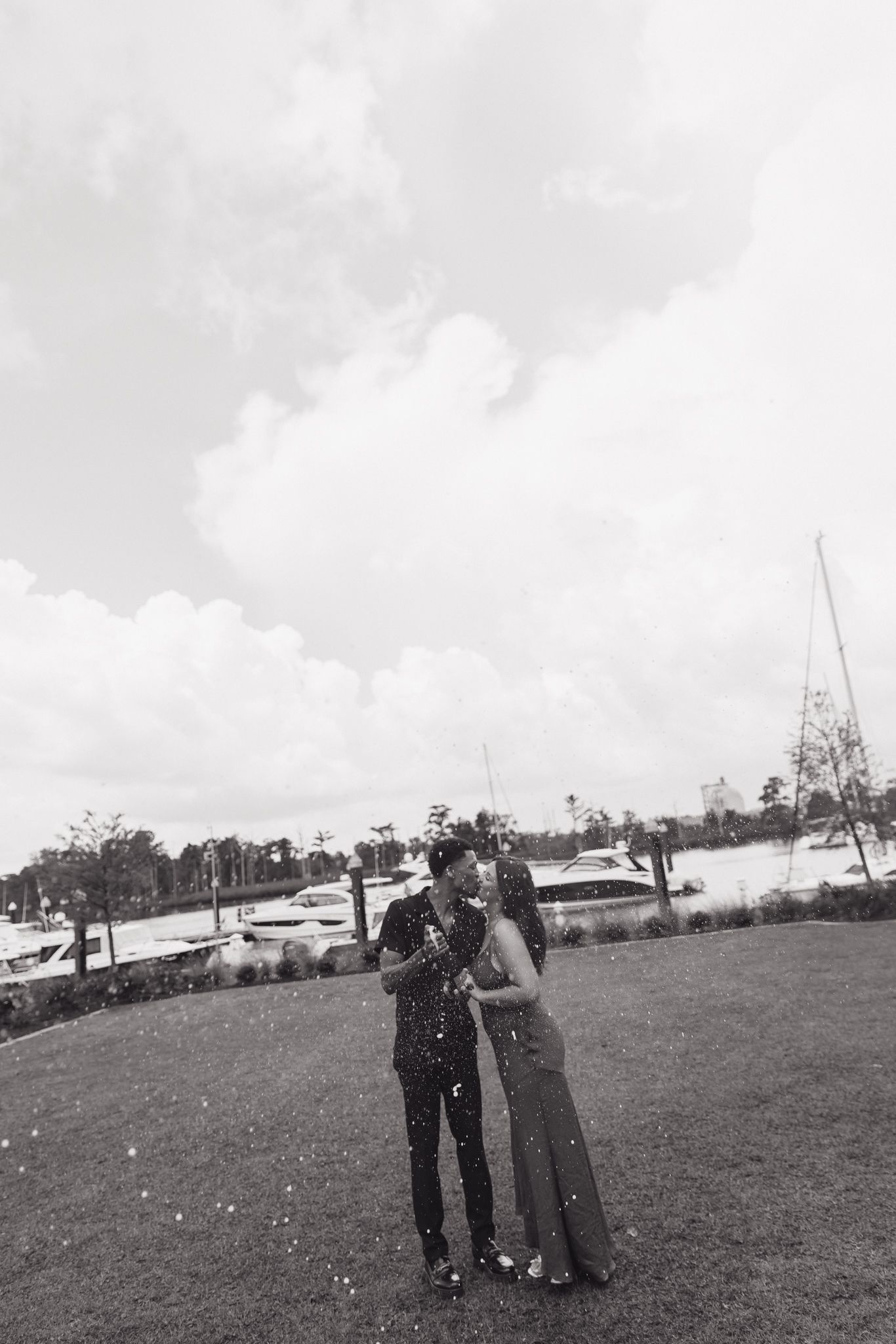 Black and white photo of couple embracing on grassy lawn with cloudy sky.