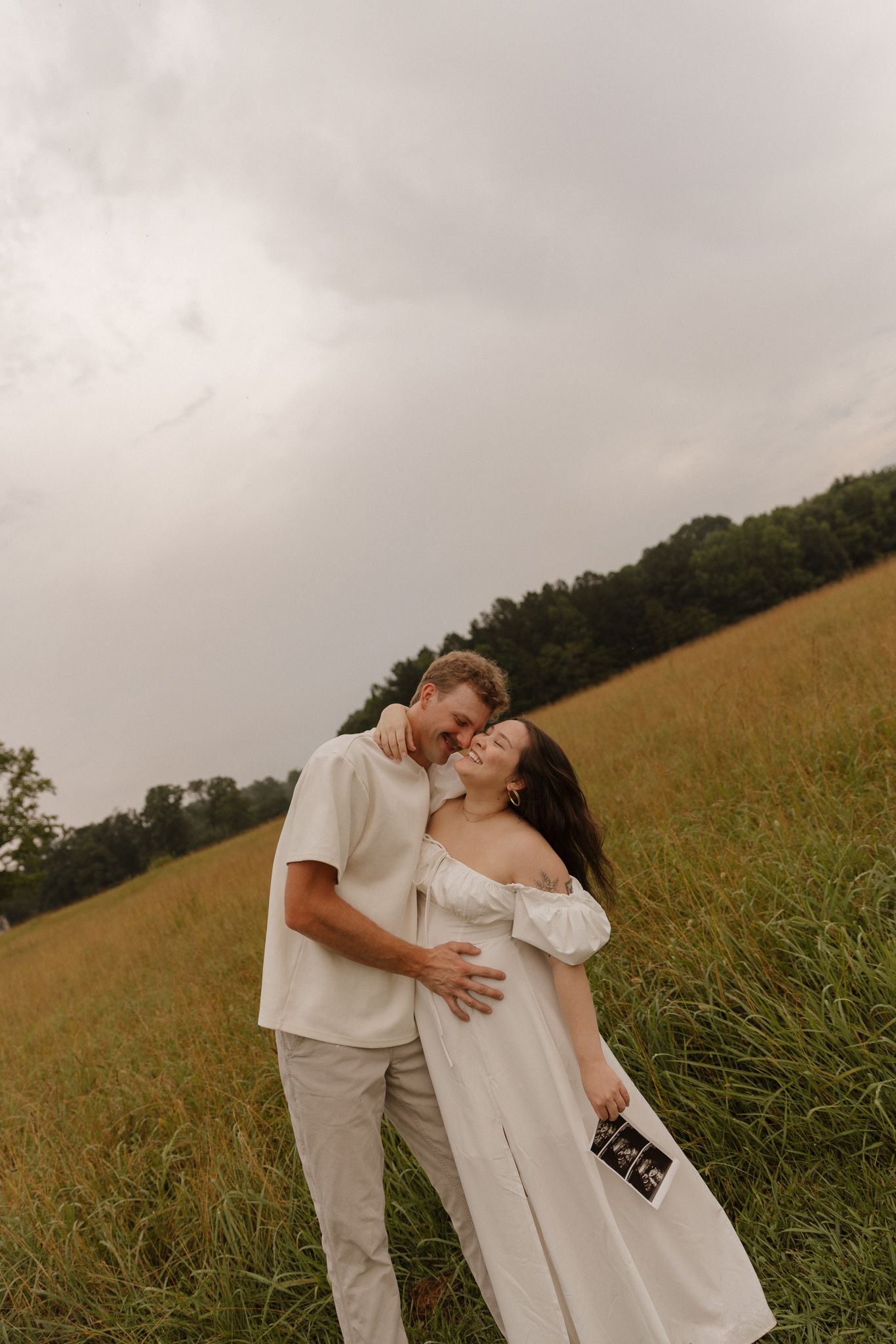 A couple embraces in a golden field under overcast skies wearing coordinating white and beige outfits.