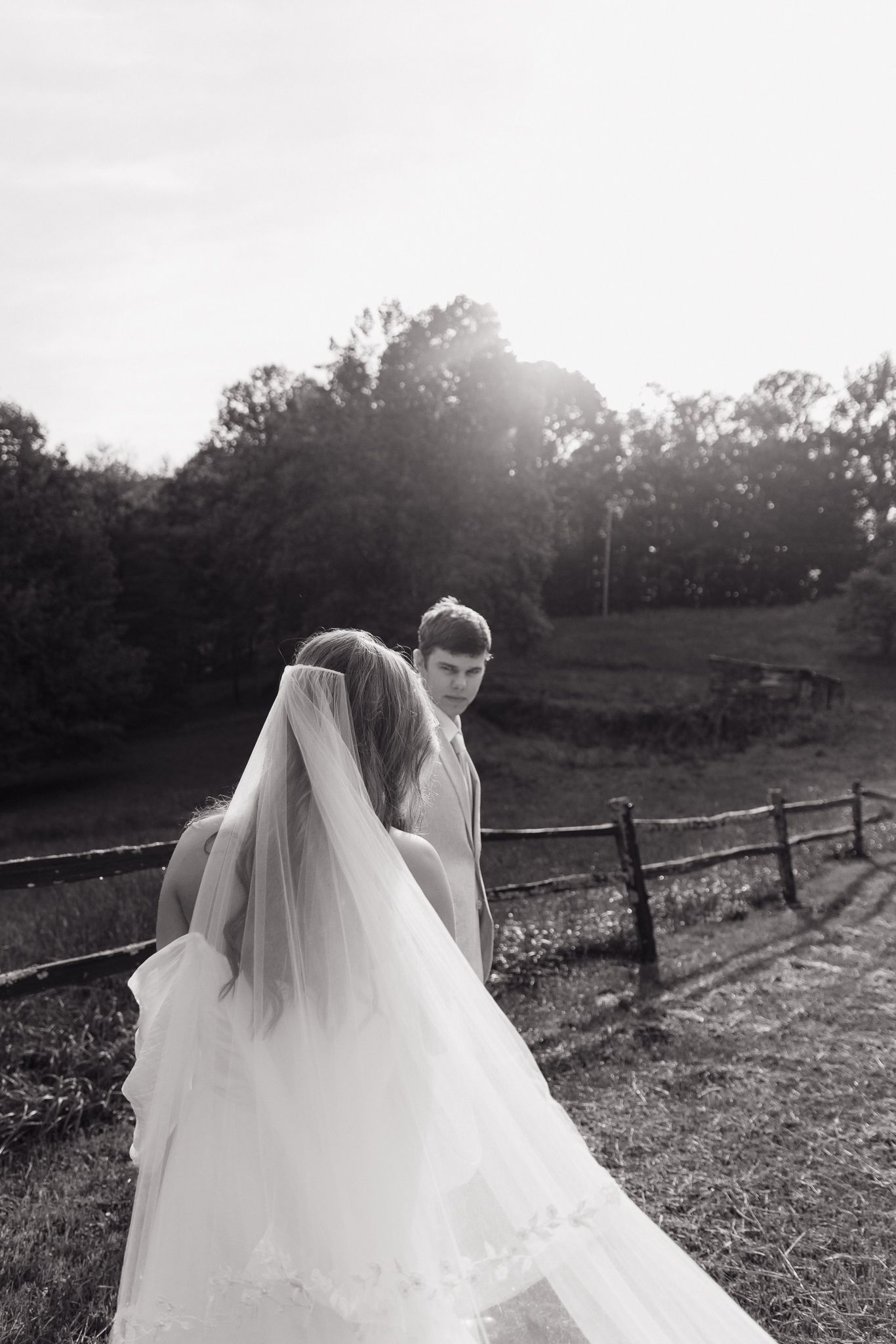 Black and white wedding portrait with flowing veil against rustic fence and trees.