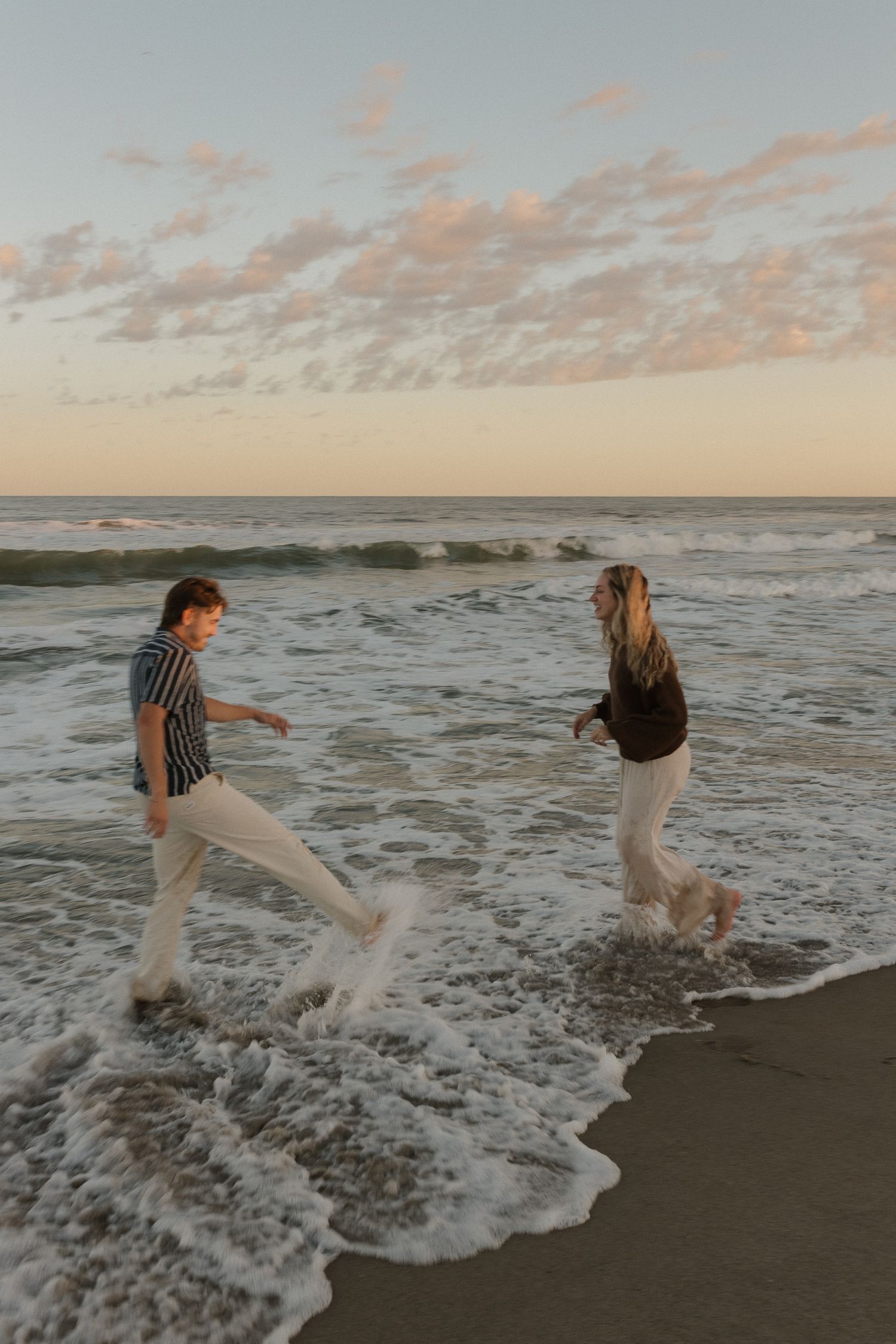 Two people playfully run and splash in the ocean waves at sunset along a sandy beach.