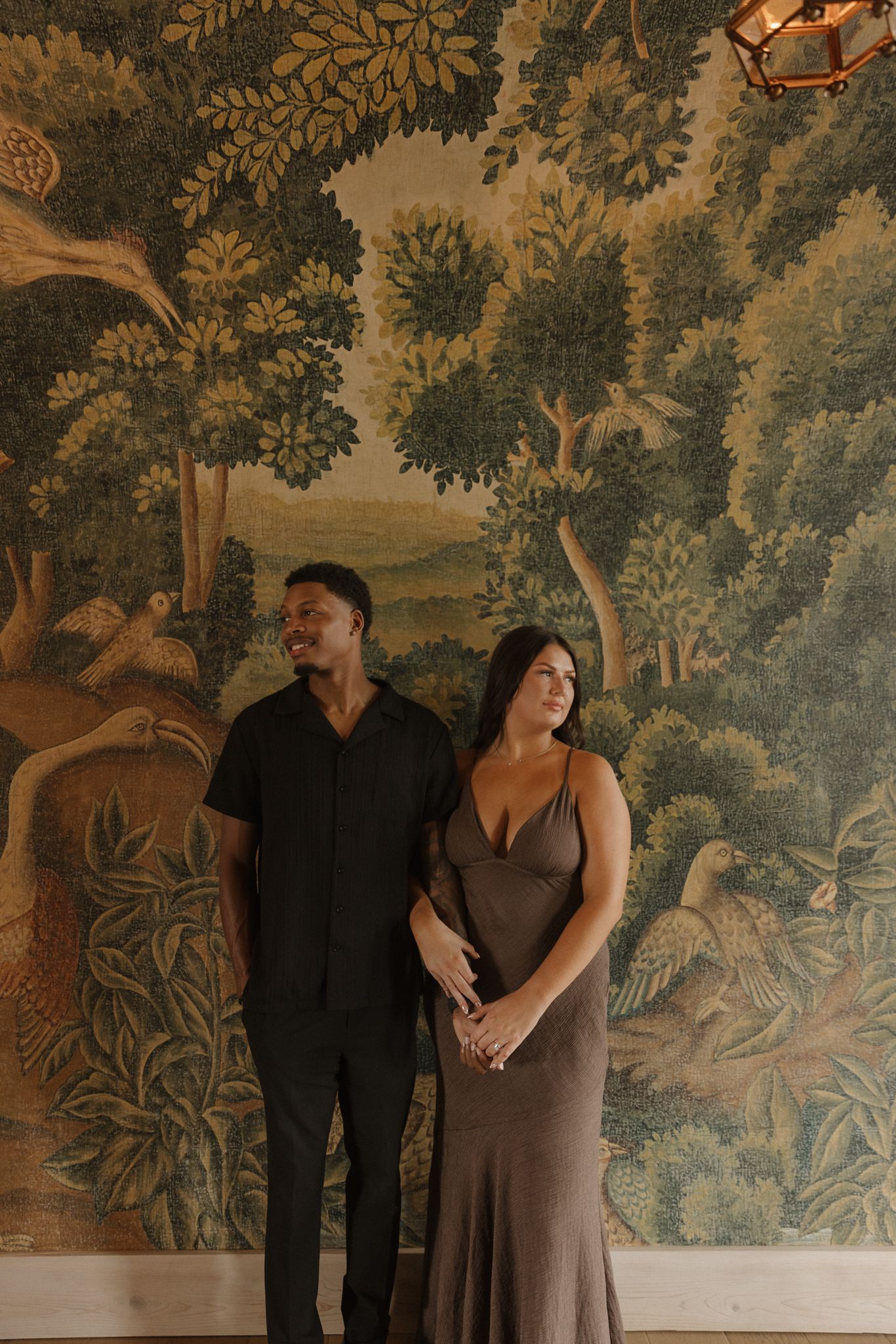 A stylish couple poses against an ornate forest wallpaper mural in formal attire.