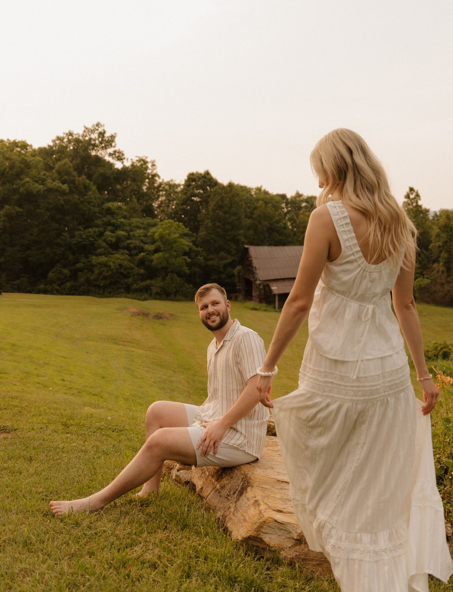 A couple sits together on a log in a grassy field at sunset, with her wearing a white tiered dress.