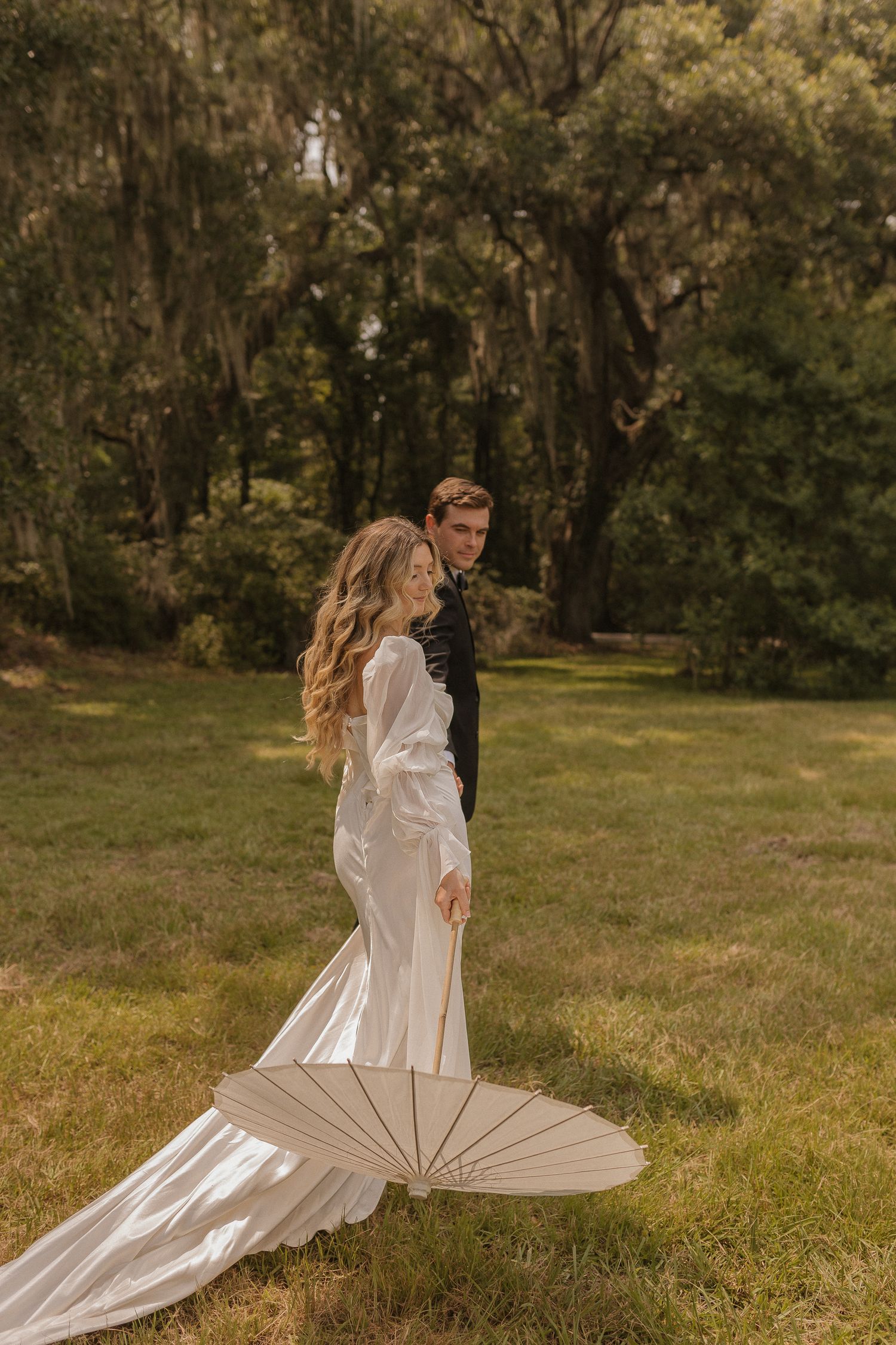 A couple poses in a moss-draped forest setting with the bride wearing a flowing white gown and holding a white parasol.