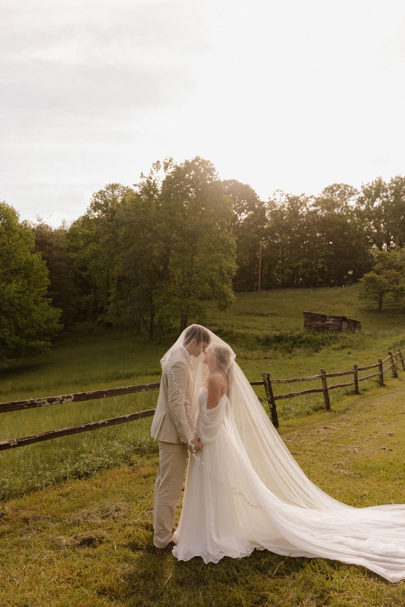 Wedding couple embraces on rolling hillside with long flowing veil and wooden fence in background.
