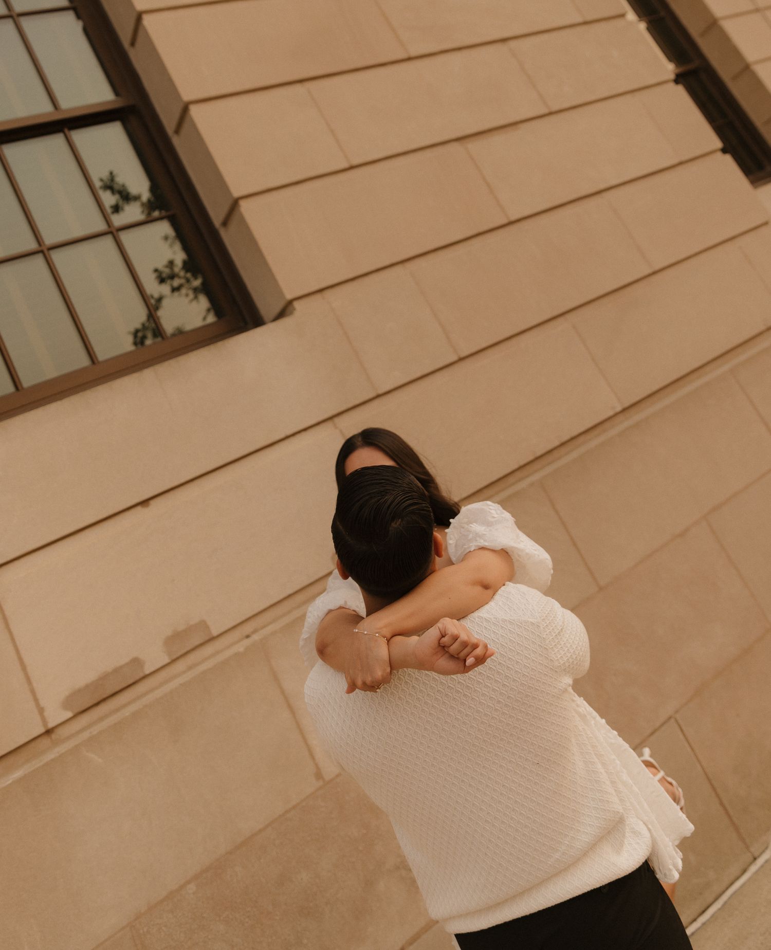 A couple embraces in warm sunlight against a beige wall with windows.
