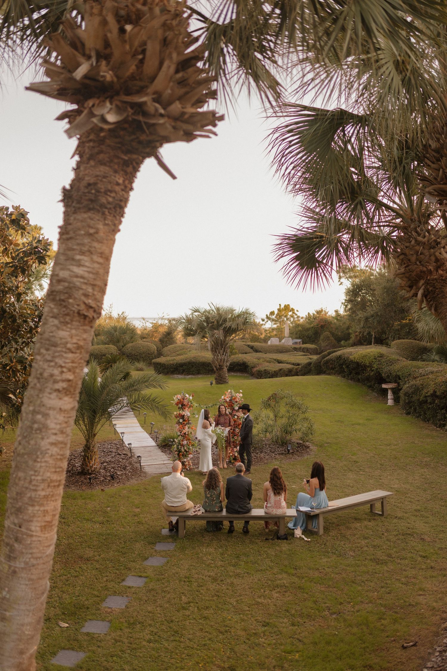 Small outdoor gathering in a garden setting with palm trees and winding pathway at sunset.