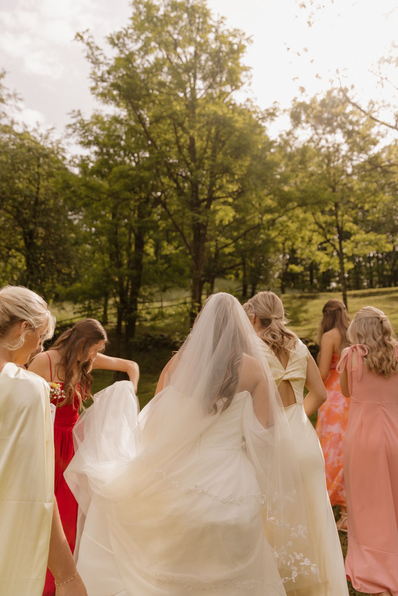 Wedding party members adjusting bridal veil in outdoor setting with trees.