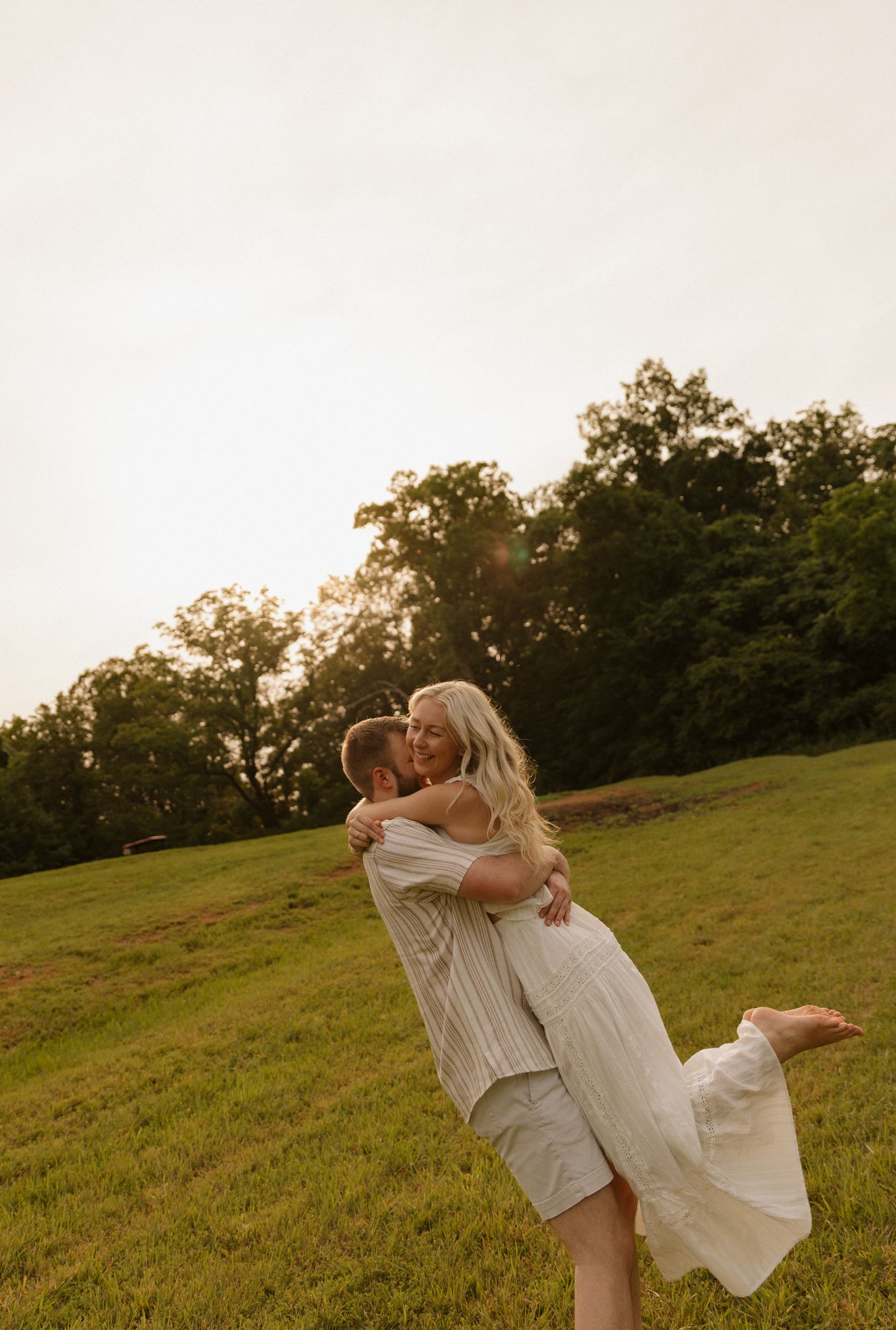 Couple in white outfits dancing together on a grassy hillside at sunset.