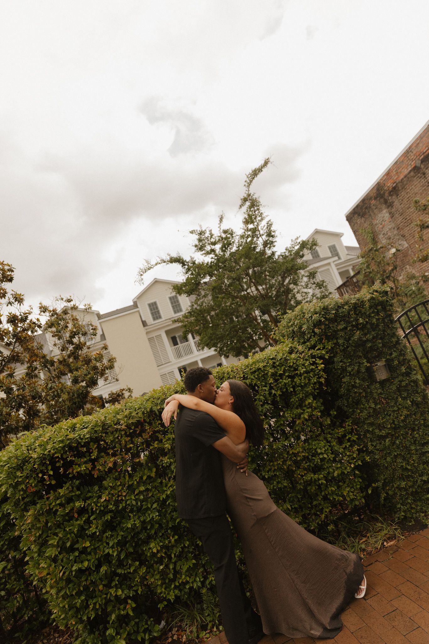 Couple sharing romantic embrace near hedges outside building.