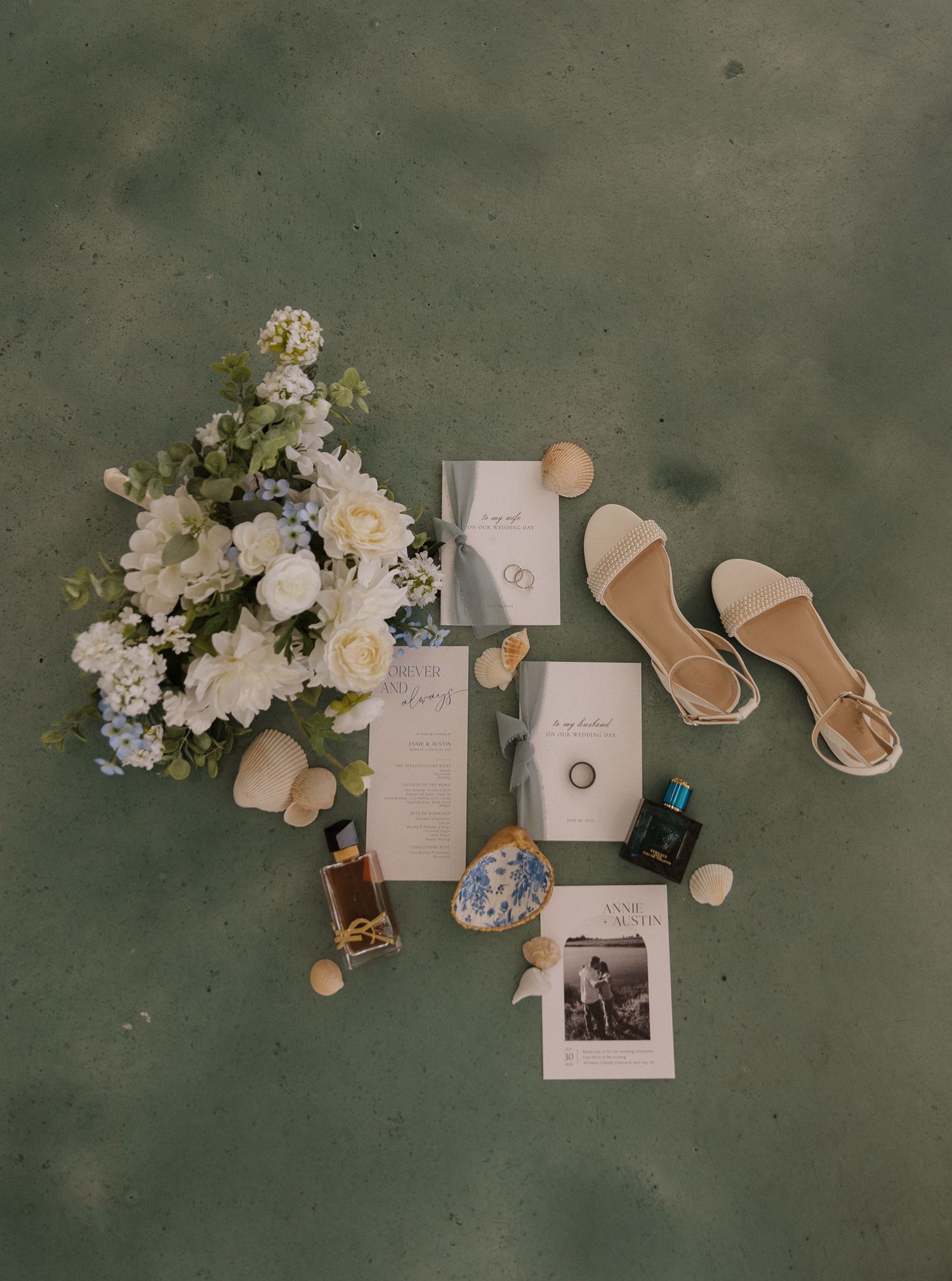 Wedding details flatlay with floral bouquet, invitation suite, jewelry and sandals on green background.