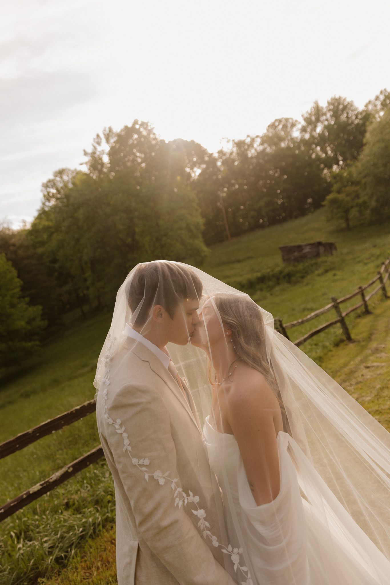 Sunset wedding portrait with couple under veil against rustic countryside backdrop.