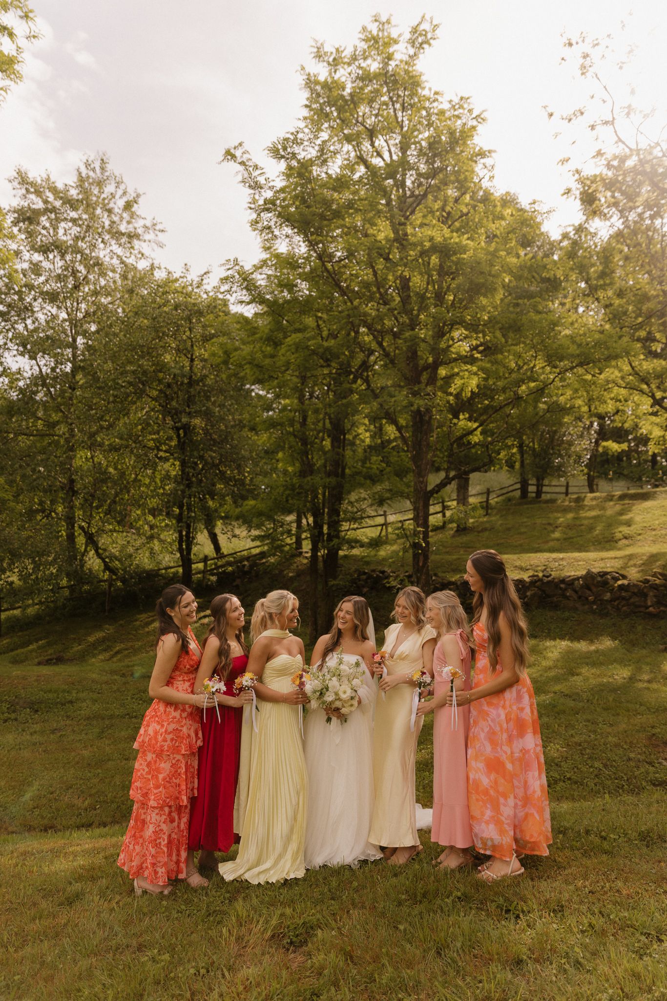 Bridal party in coral and pink dresses posing outdoors against autumn trees.