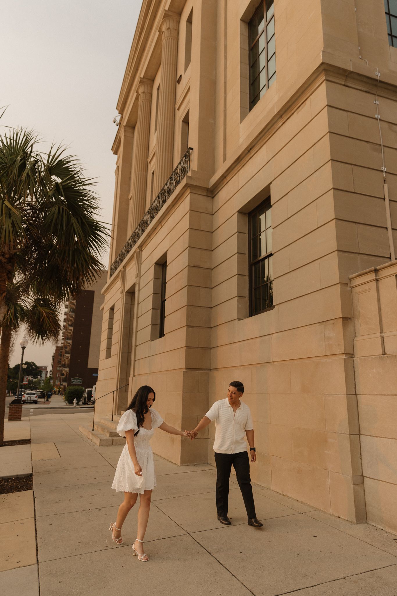 A couple holds hands while walking along a historic building facade with palm trees.