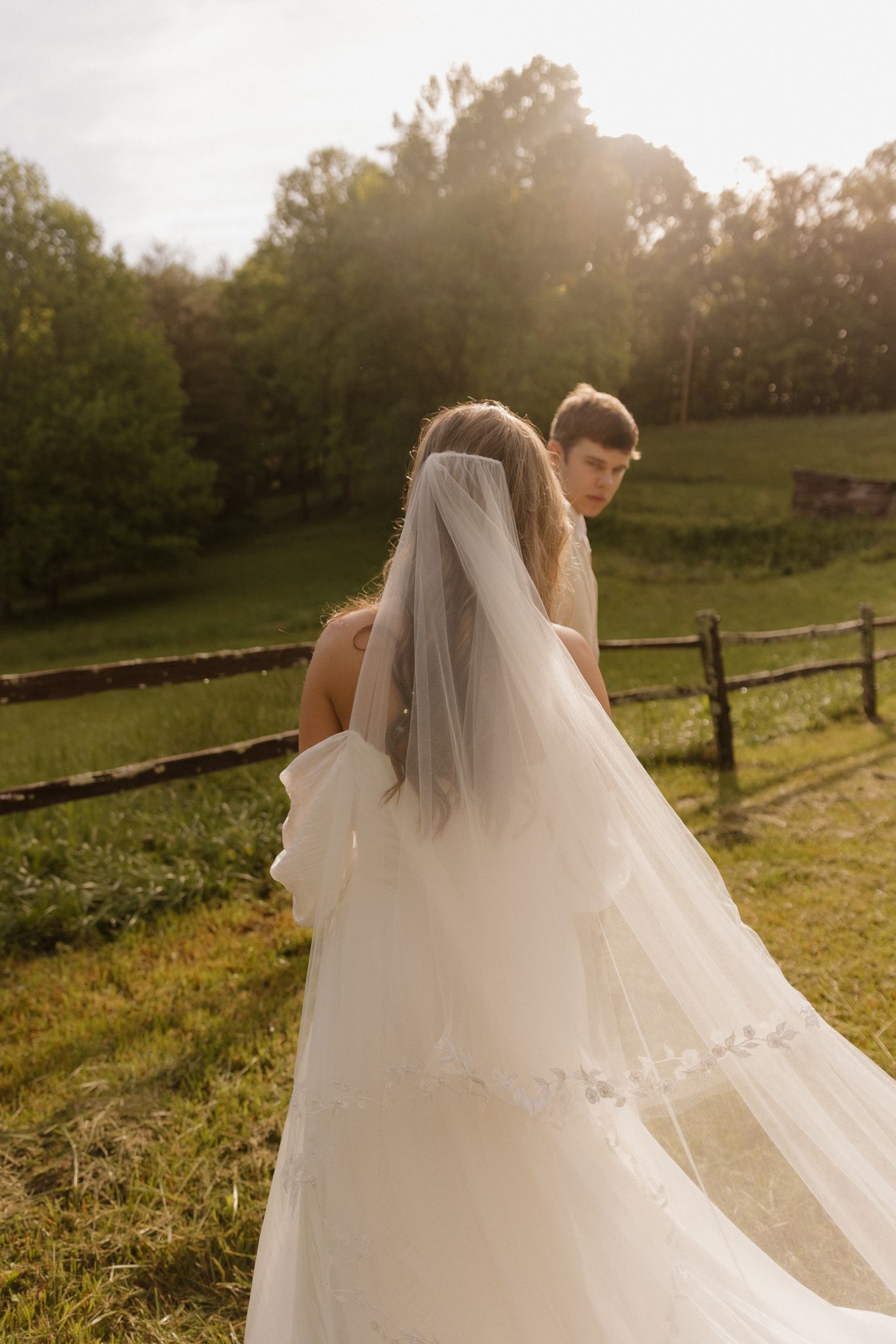 Candid wedding moment captured in golden hour light with flowing veil and rustic fence.