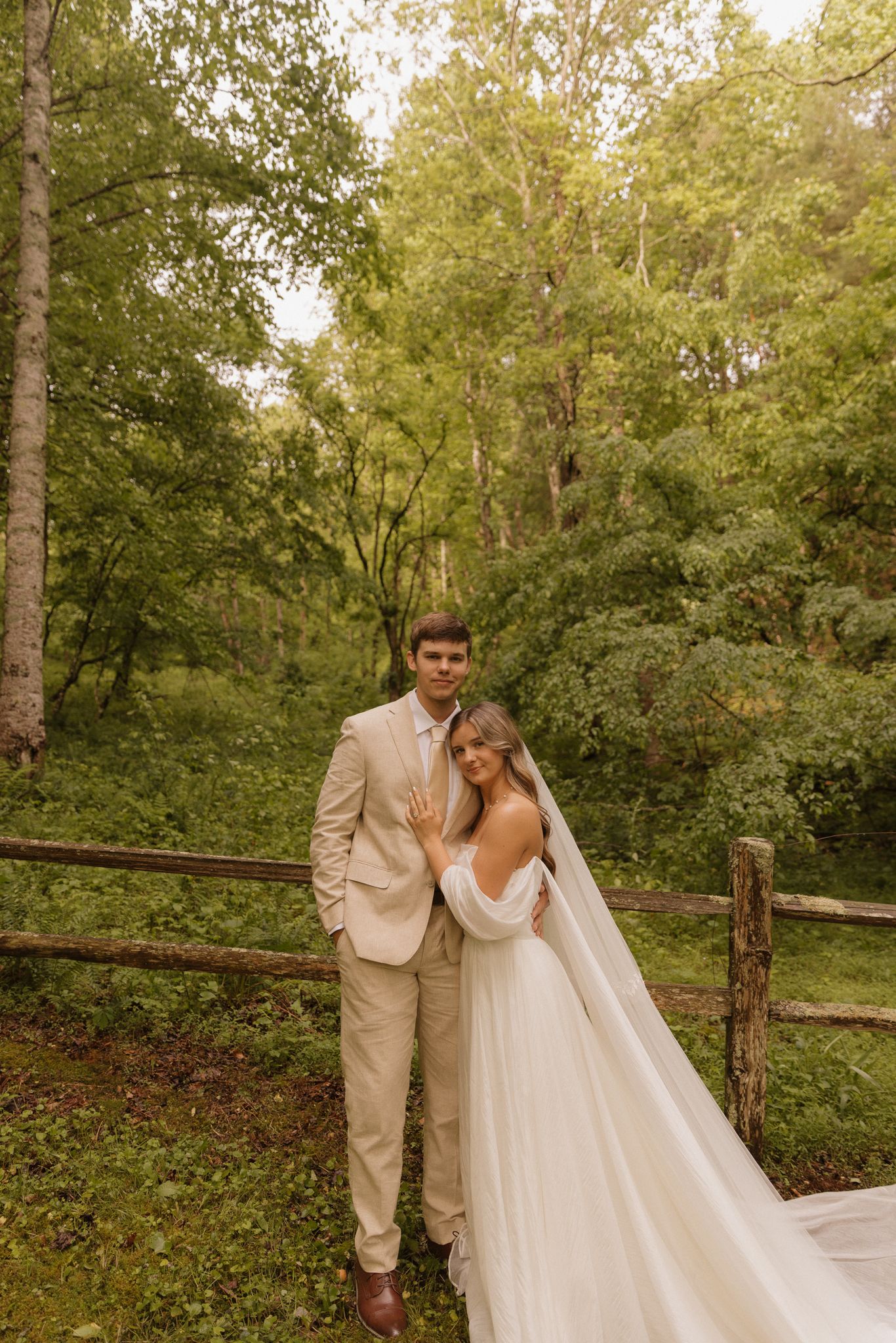 Couple poses together in a natural woodland setting with rustic fence, wearing elegant light-colored wedding attire.