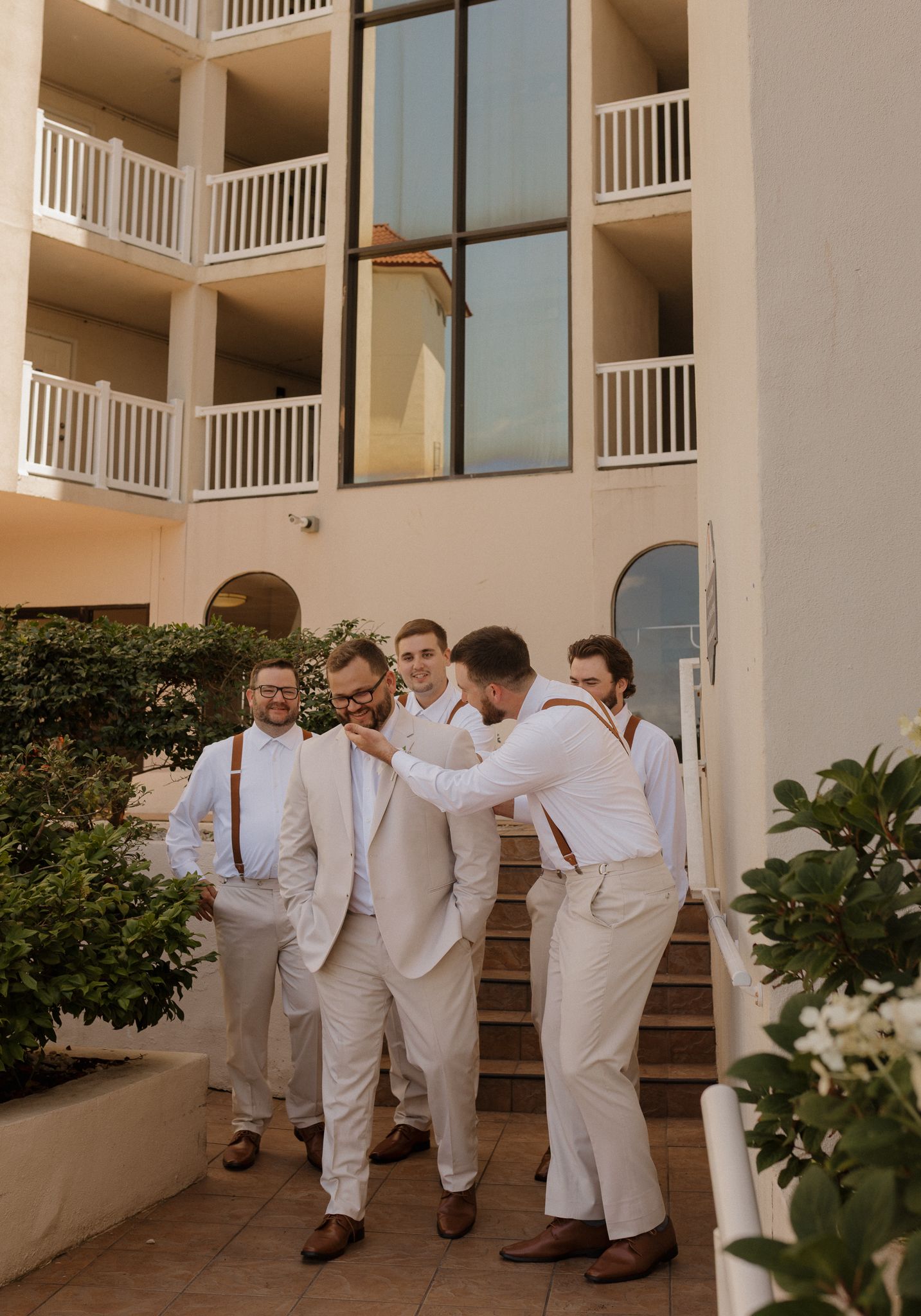 Groomsmen in white suits walking and laughing together outside a beachfront building.