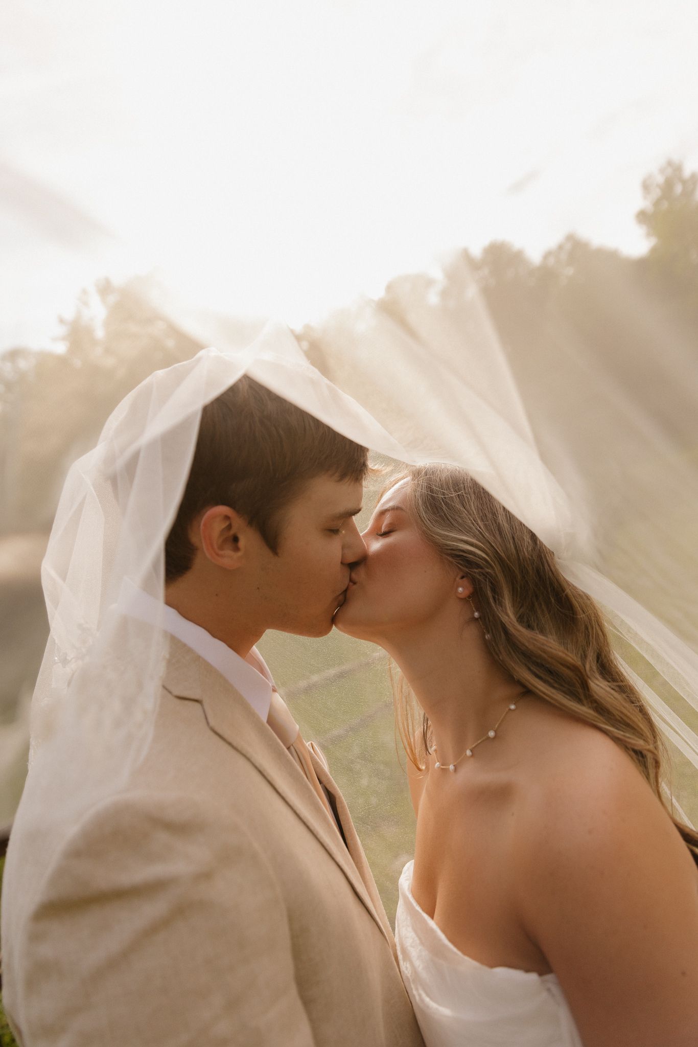 Romantic sunset kiss captured between newlyweds with flowing veil in golden light.