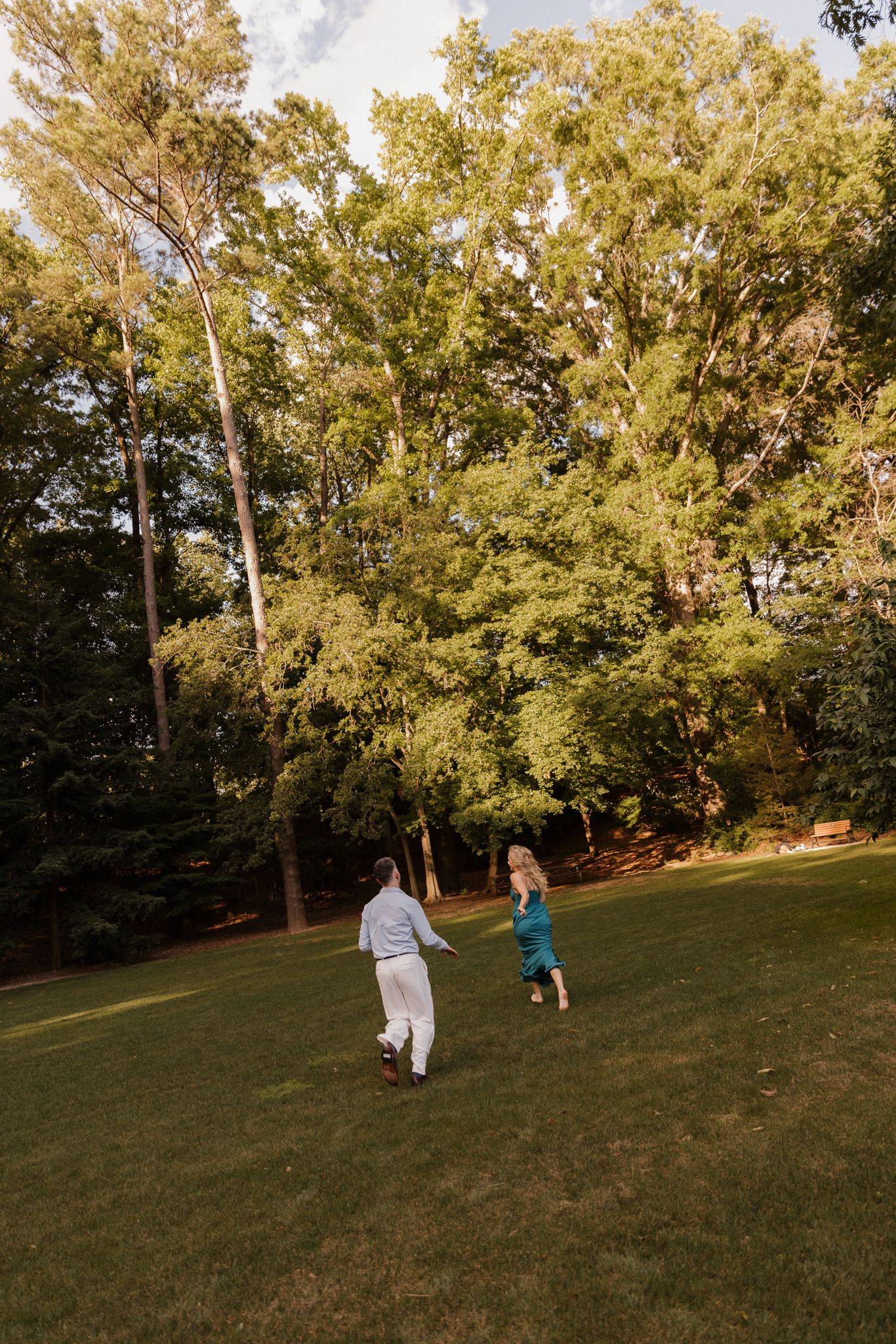 Two people running joyfully across a sunlit lawn surrounded by tall trees.