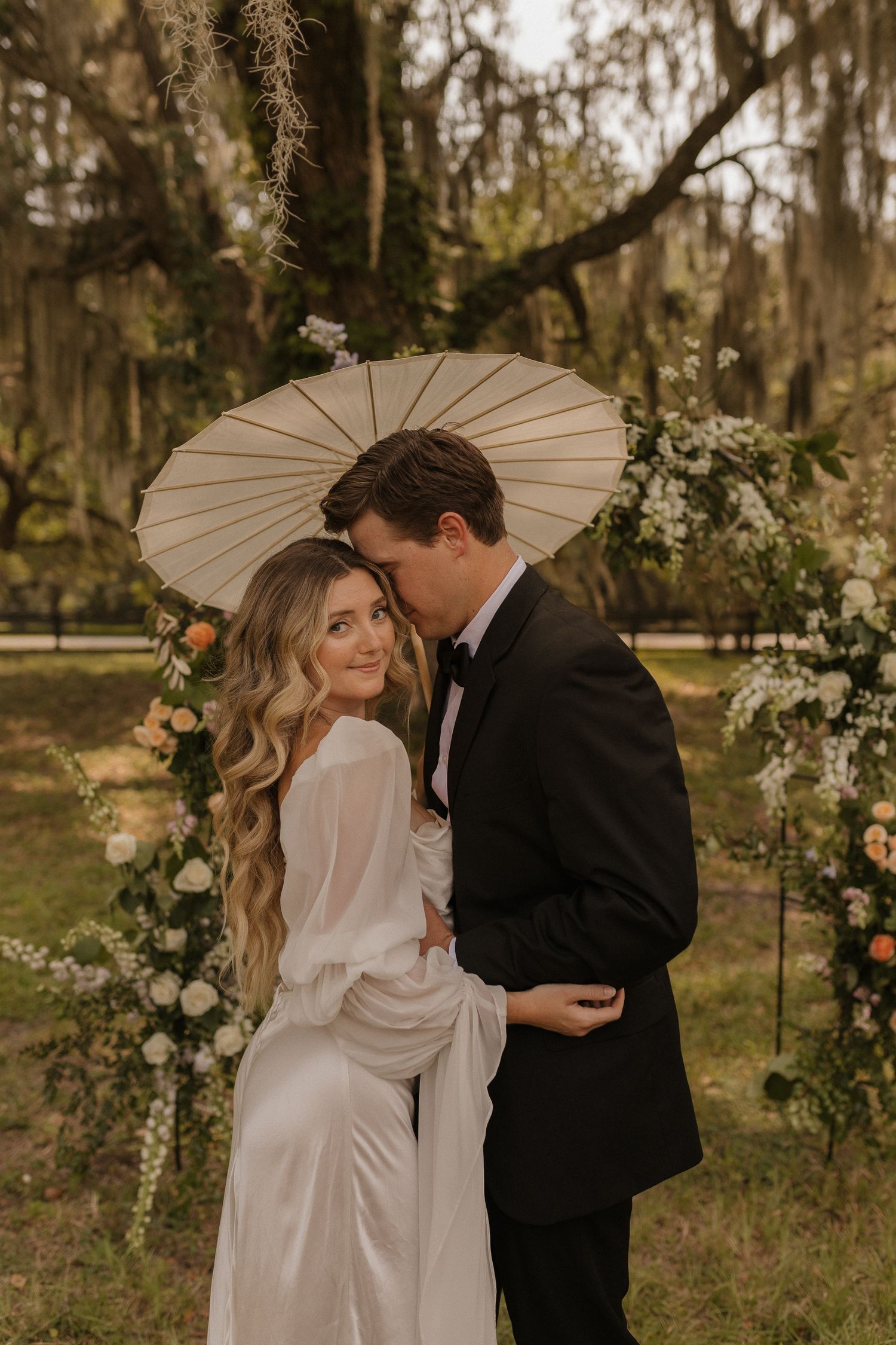 A romantic wedding couple shares an intimate moment under a white parasol in a Spanish moss-draped garden setting.