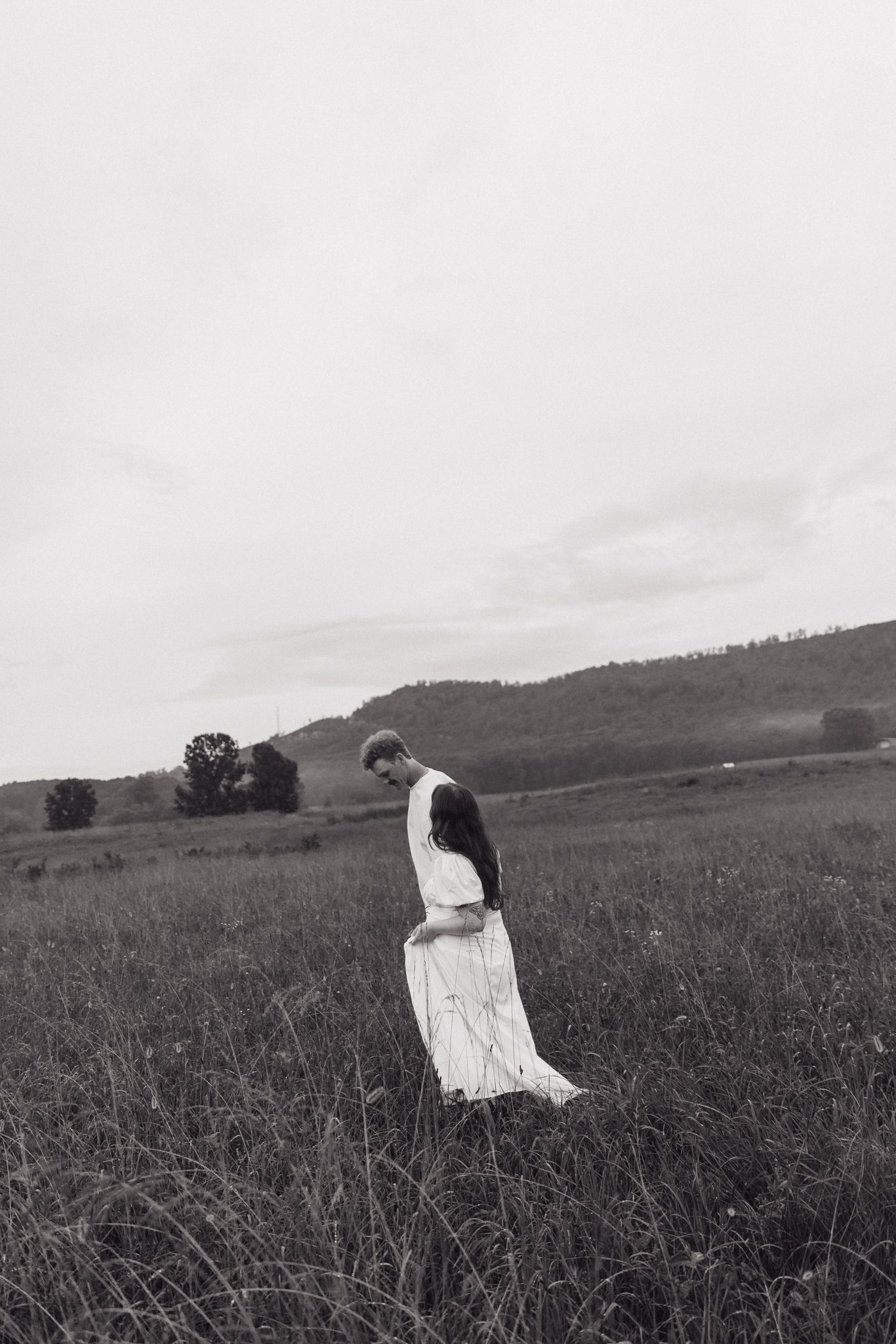 Black and white photograph of a figure in white dress crouching in a field with mountains in the distance.