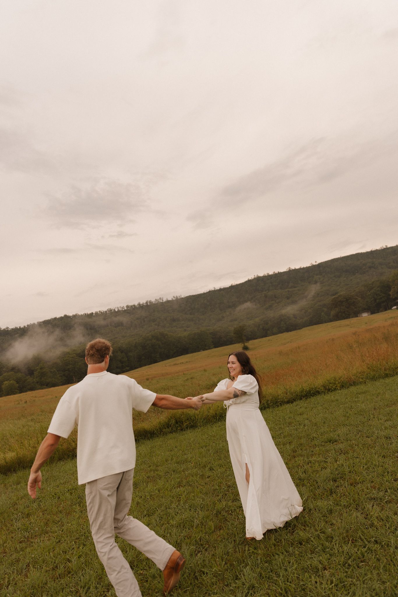 Two people in white outfits dance freely across a scenic mountain meadow under cloudy skies.