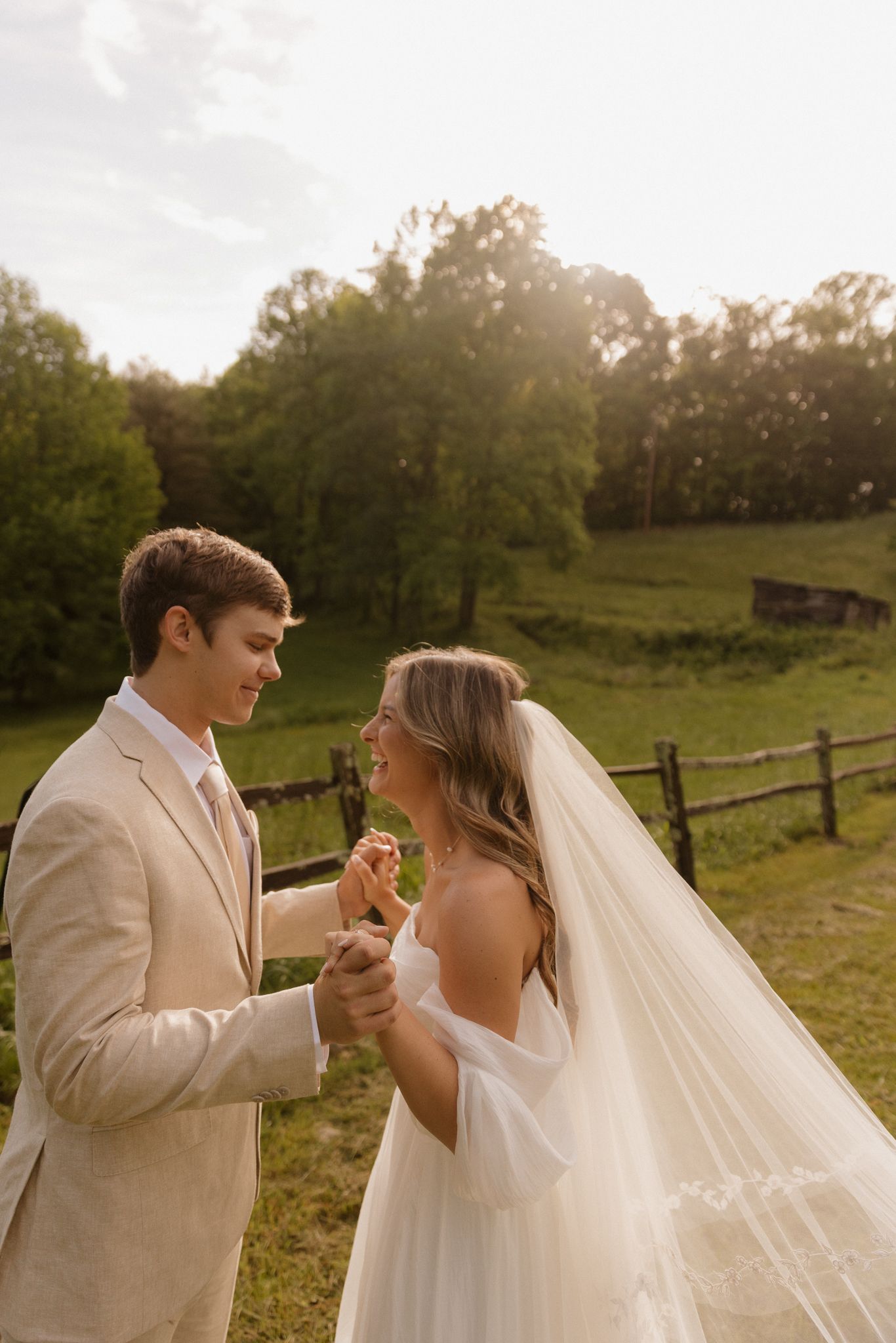 Newlyweds share a romantic moment in a pastoral setting with rustic fencing and golden sunlight.