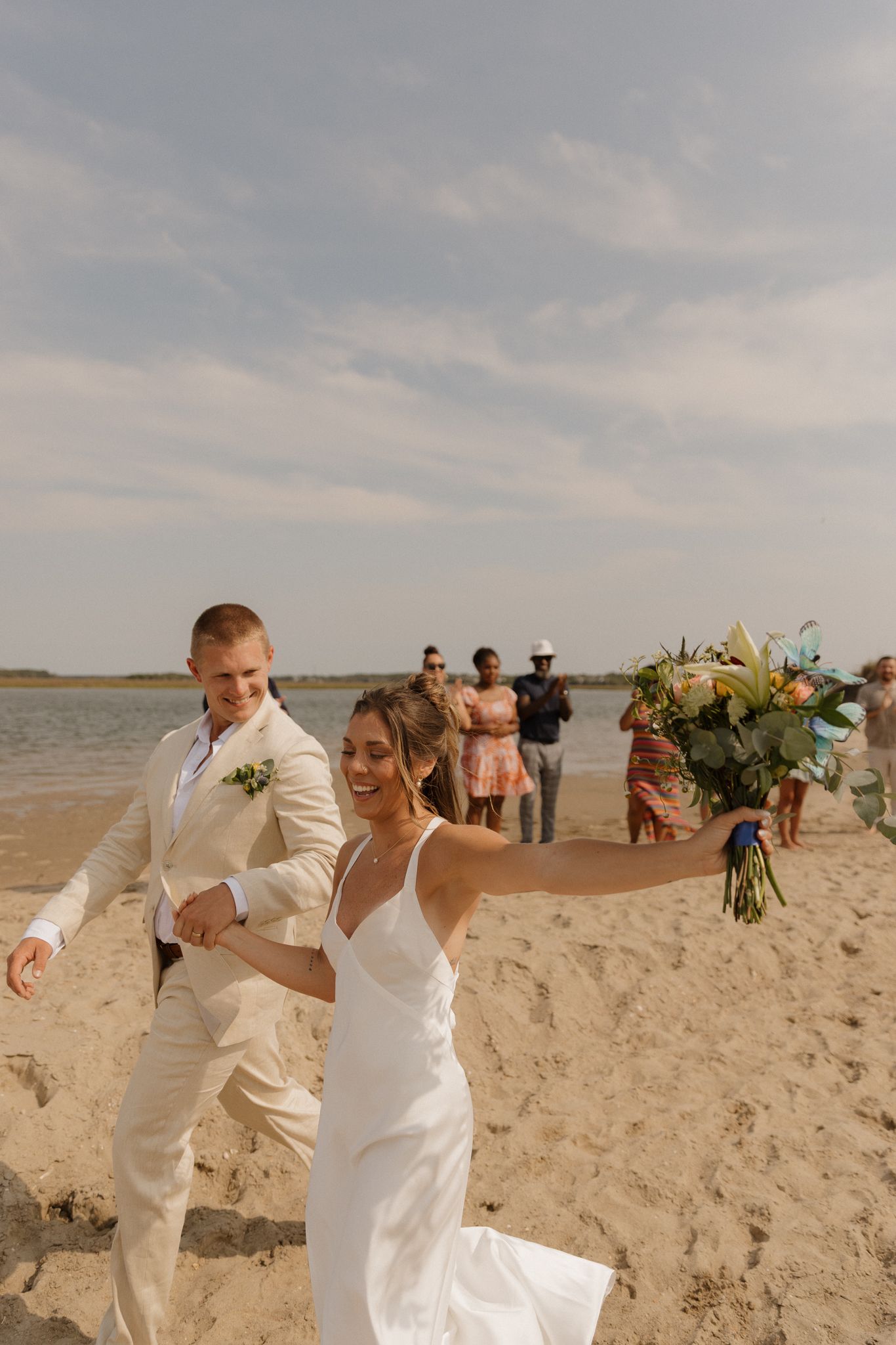 Newlyweds dancing joyfully on a beach with bouquets of yellow flowers and white attire.