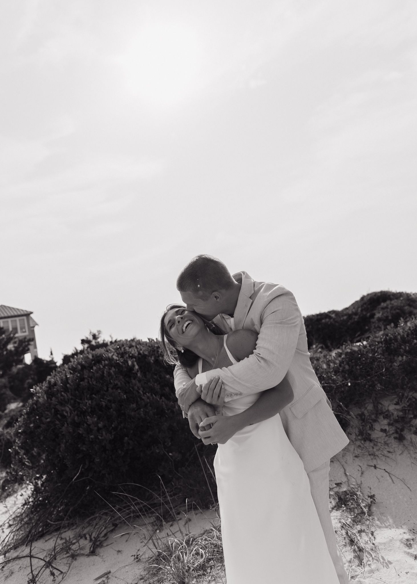 A romantic black and white beach wedding photo with a couple embracing against coastal vegetation.