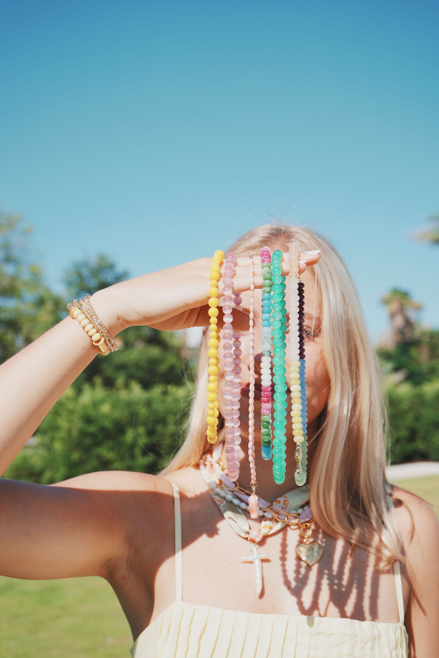 Colorful beaded bracelets are displayed against a bright blue sky background with green foliage in the distance.