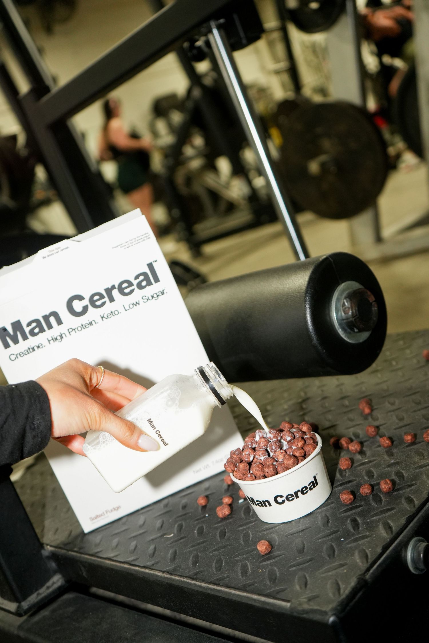 A box of Man Cereal protein cereal being poured into a bowl at a gym with exercise equipment in the background.