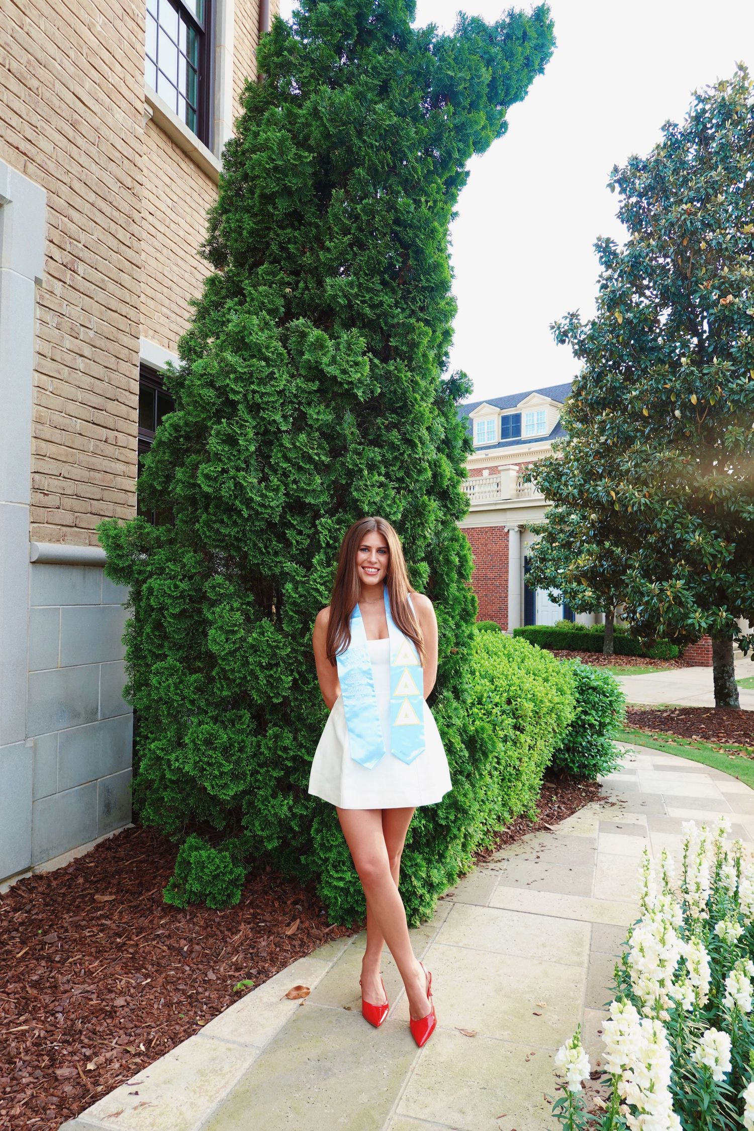 A person in a light blue and white summer dress stands on a sidewalk next to landscaping and brick buildings.