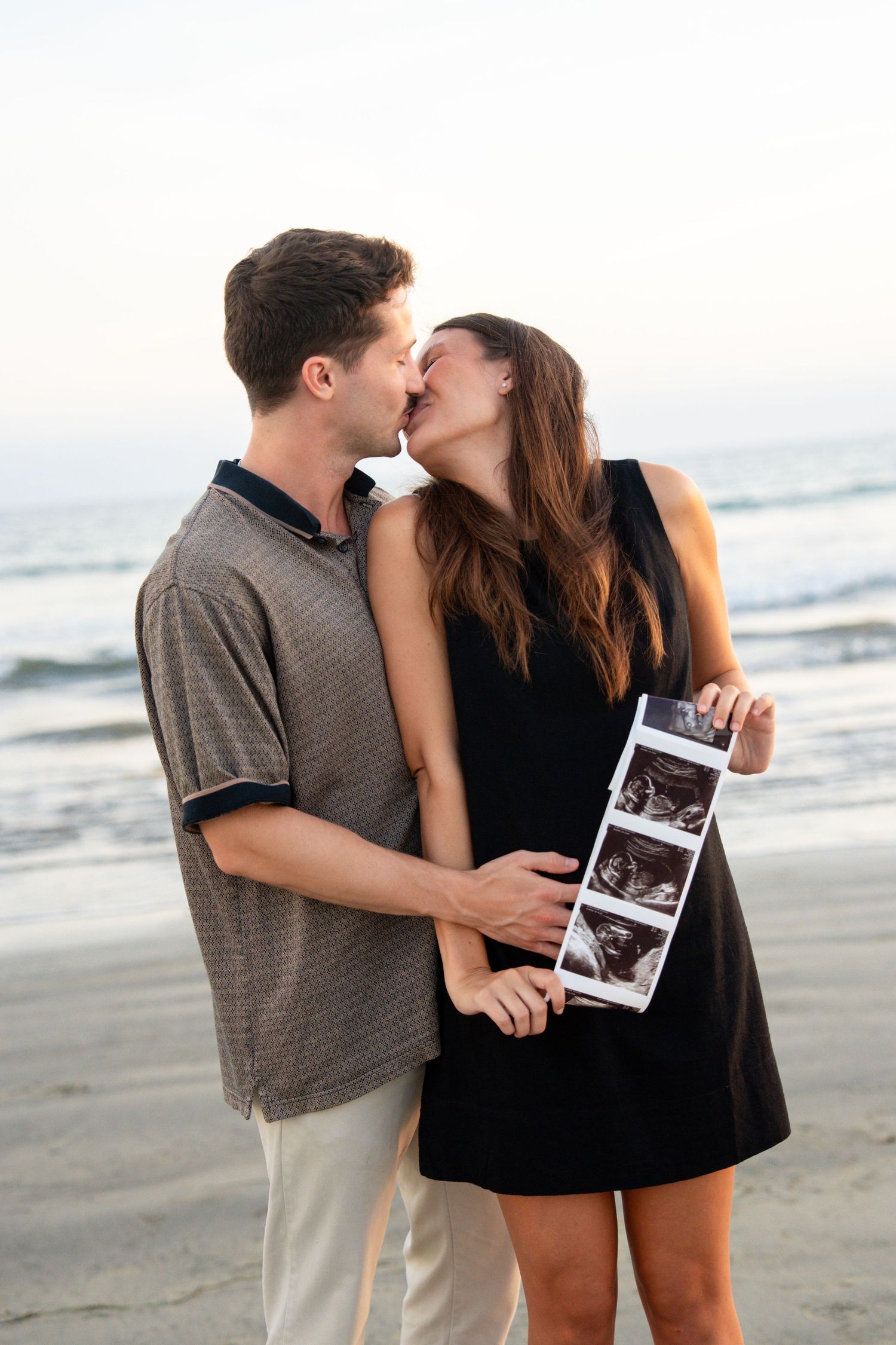 Couple kissing on beach holding ultrasound photos, announcing pregnancy near ocean waves.