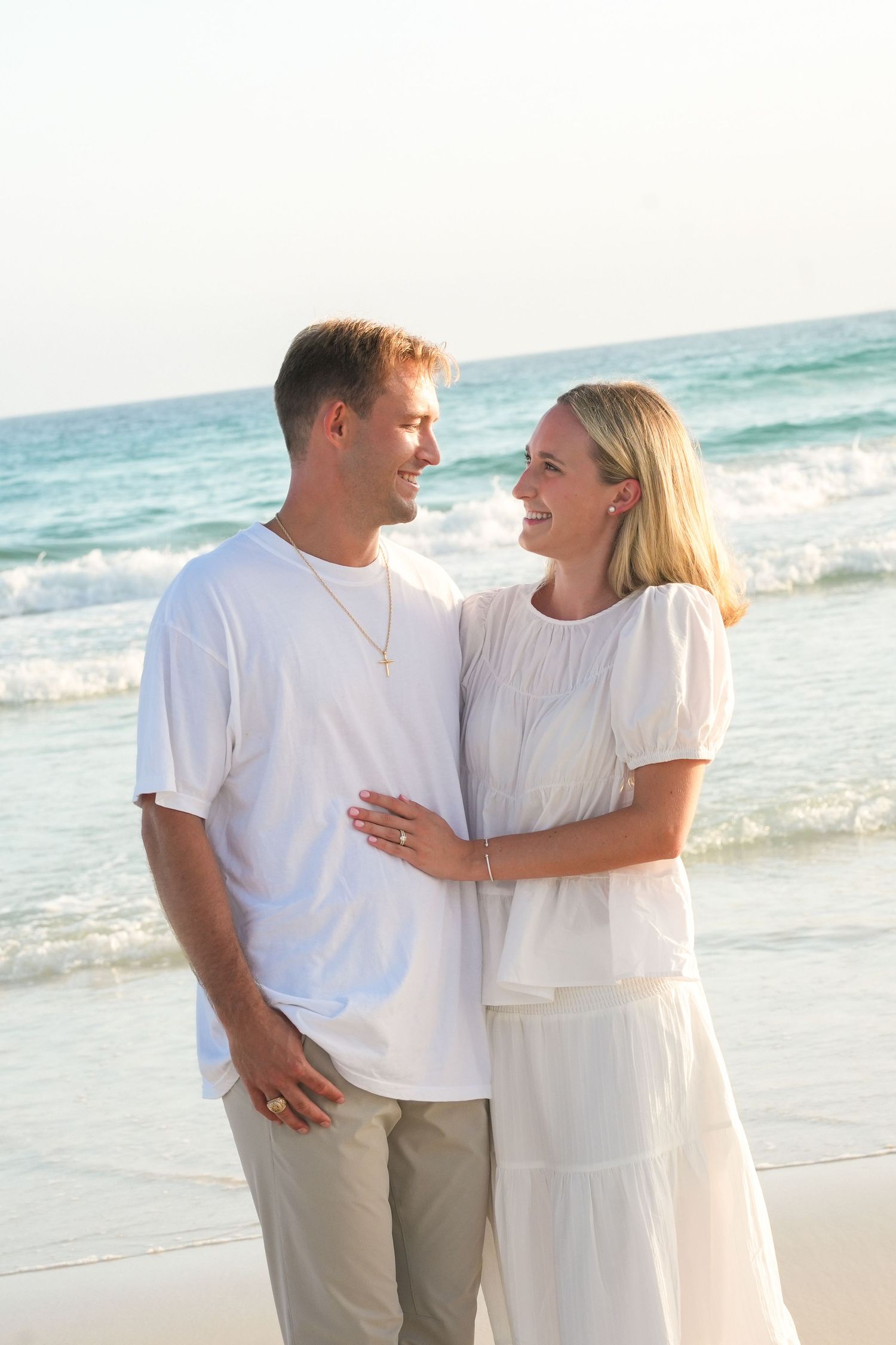 Happy couple in white outfits embracing on a sunny beach with ocean waves in the background.