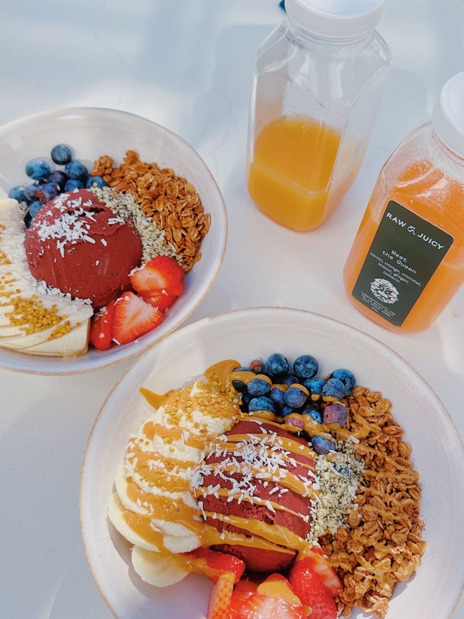 Colorful breakfast bowls with acai, granola, fresh berries and juice bottles on white surface.