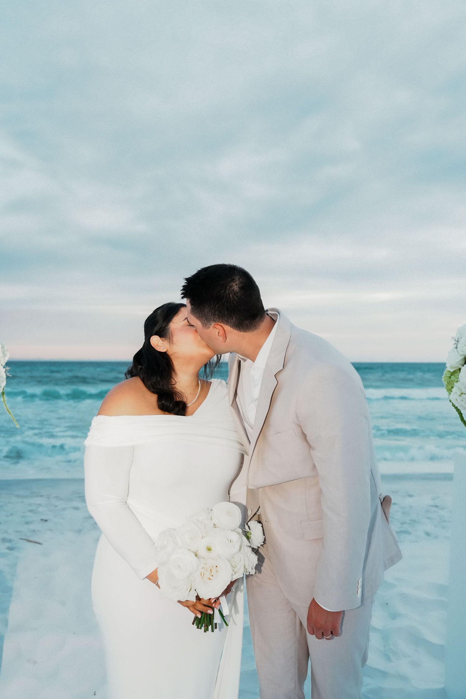 A romantic moment captured during a beach wedding with white florals and gentle ocean waves in the background.