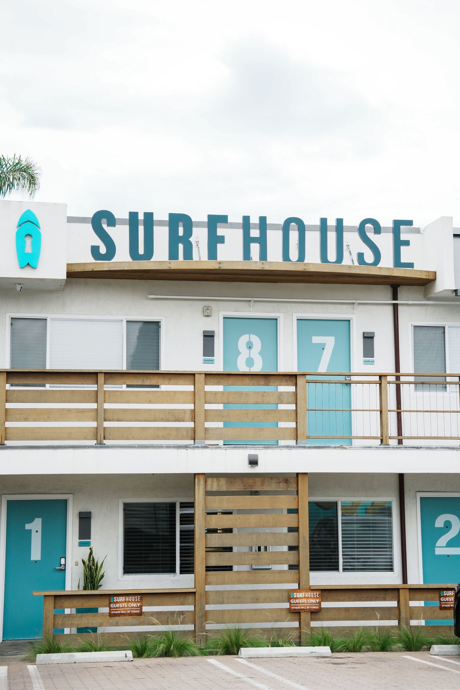 White and turquoise surfhouse motel exterior with wooden slat railing and room numbers.