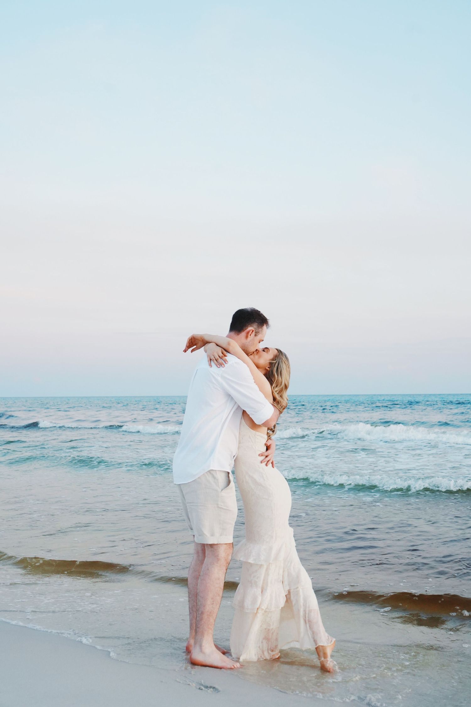 A white-clad couple shares a romantic moment at sunset along the ocean shore.