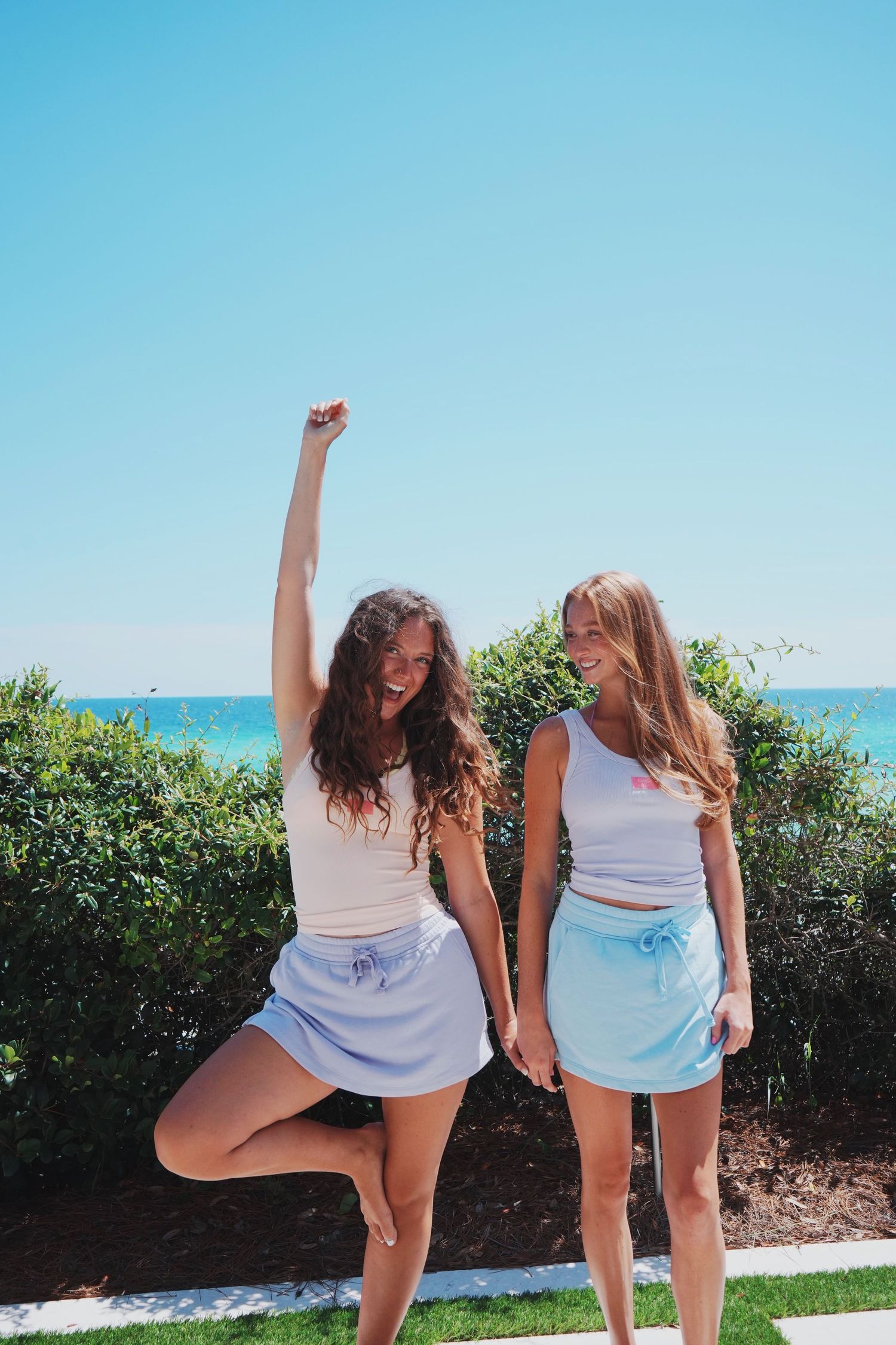Two people pose playfully in matching white outfits on a sunny day by the beach with ocean and greenery visible.
