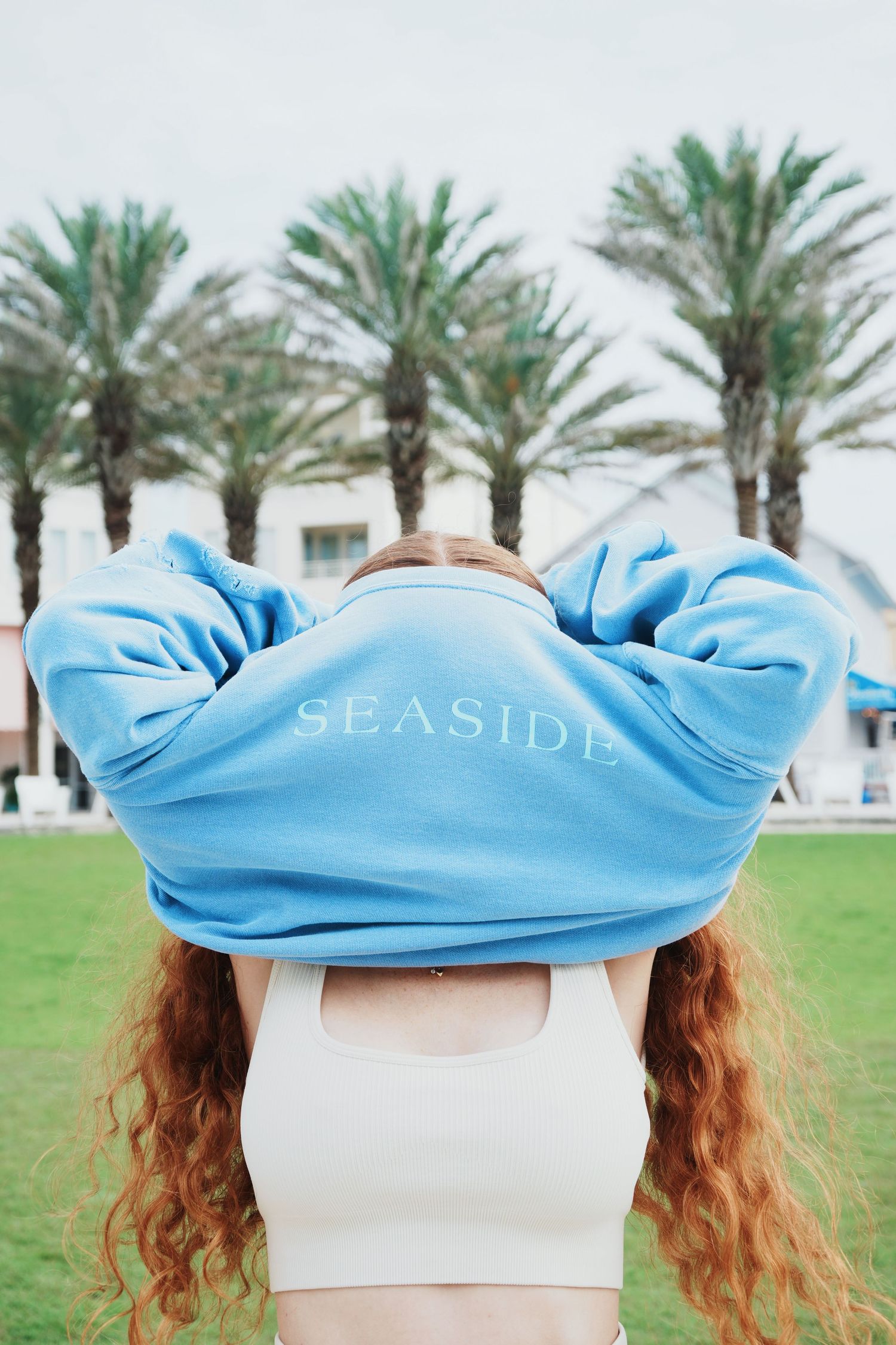 Light blue Seaside sweatshirt worn backwards against palm trees and green lawn backdrop.