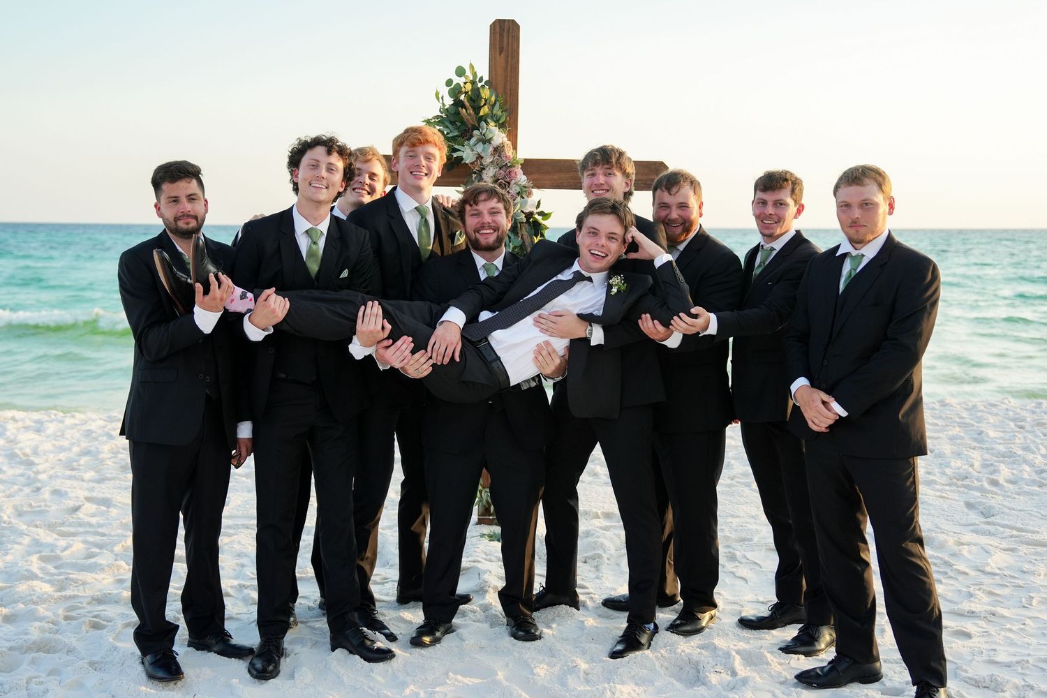 Groomsmen in black suits lifting groom on a beach wedding with a wooden cross backdrop.