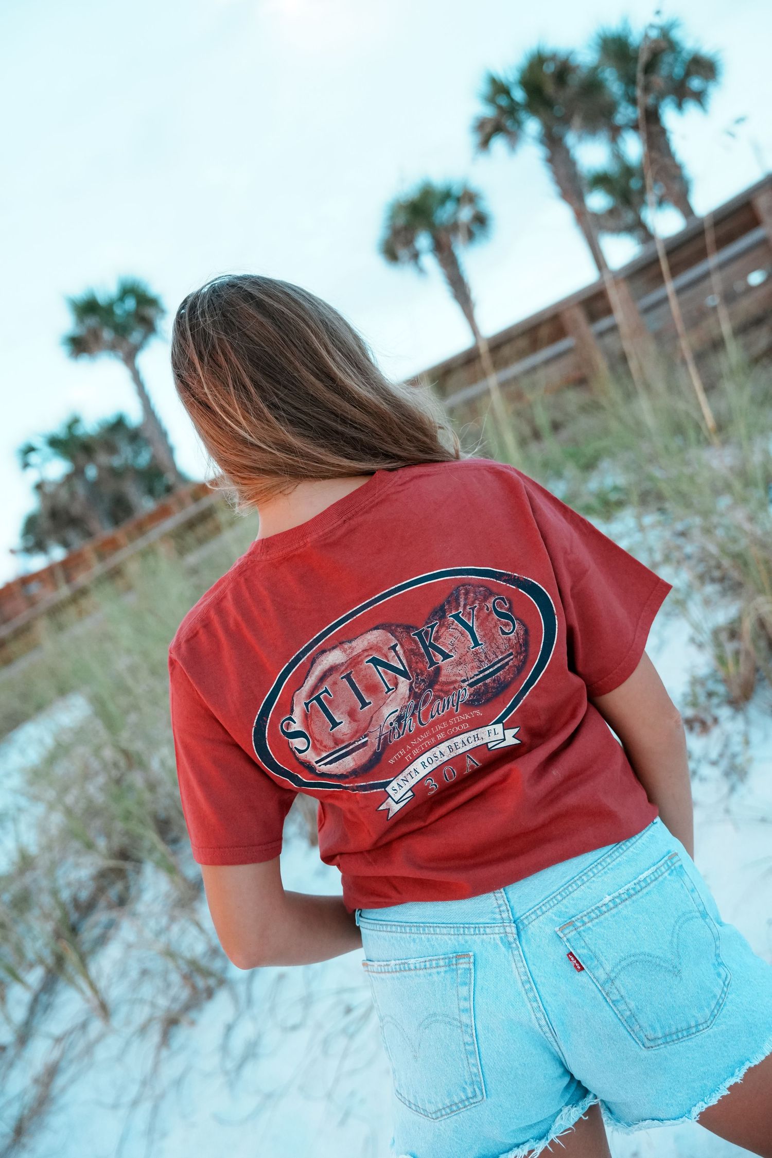 Person wearing a red t-shirt with graphic design and denim shorts poses on a beach.