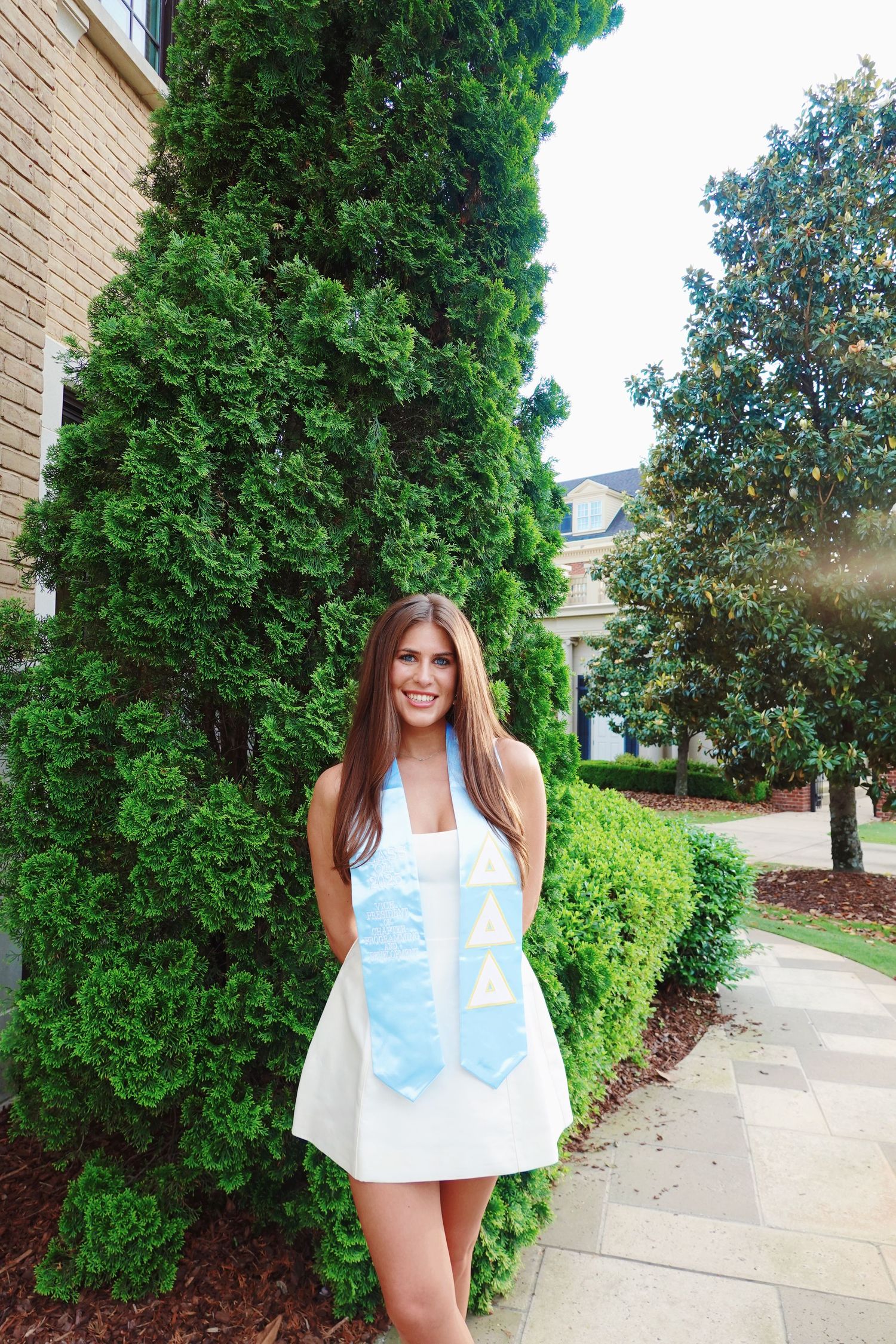 Summer fashion photo against green shrubs and brick building featuring a white halter dress.